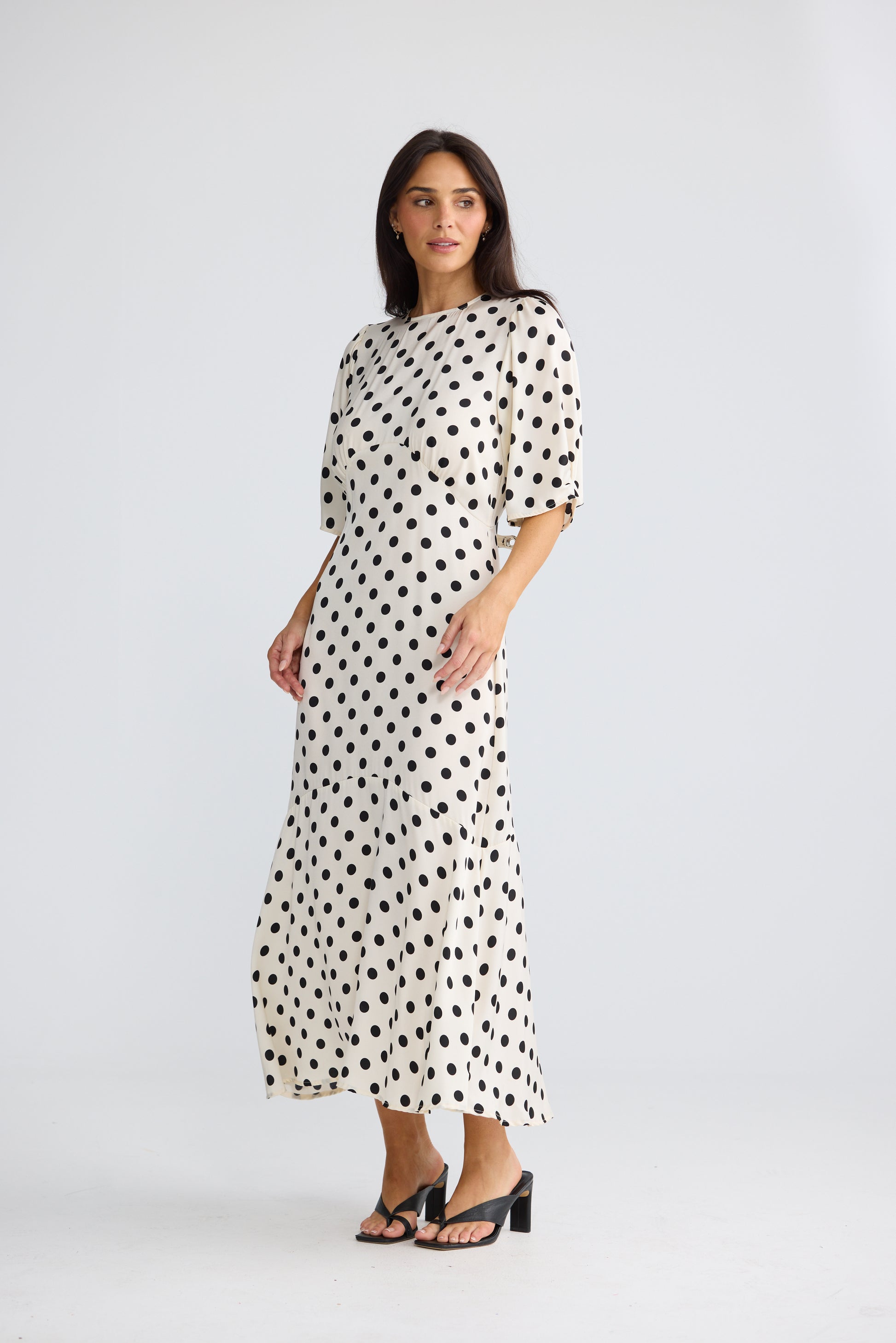 A woman modeling a Brave & True midi-length cream dress with a black polka dot pattern, featuring elbow-length puff sleeves and a high round neckline, paired with black strappy heels against a light gray studio background.
