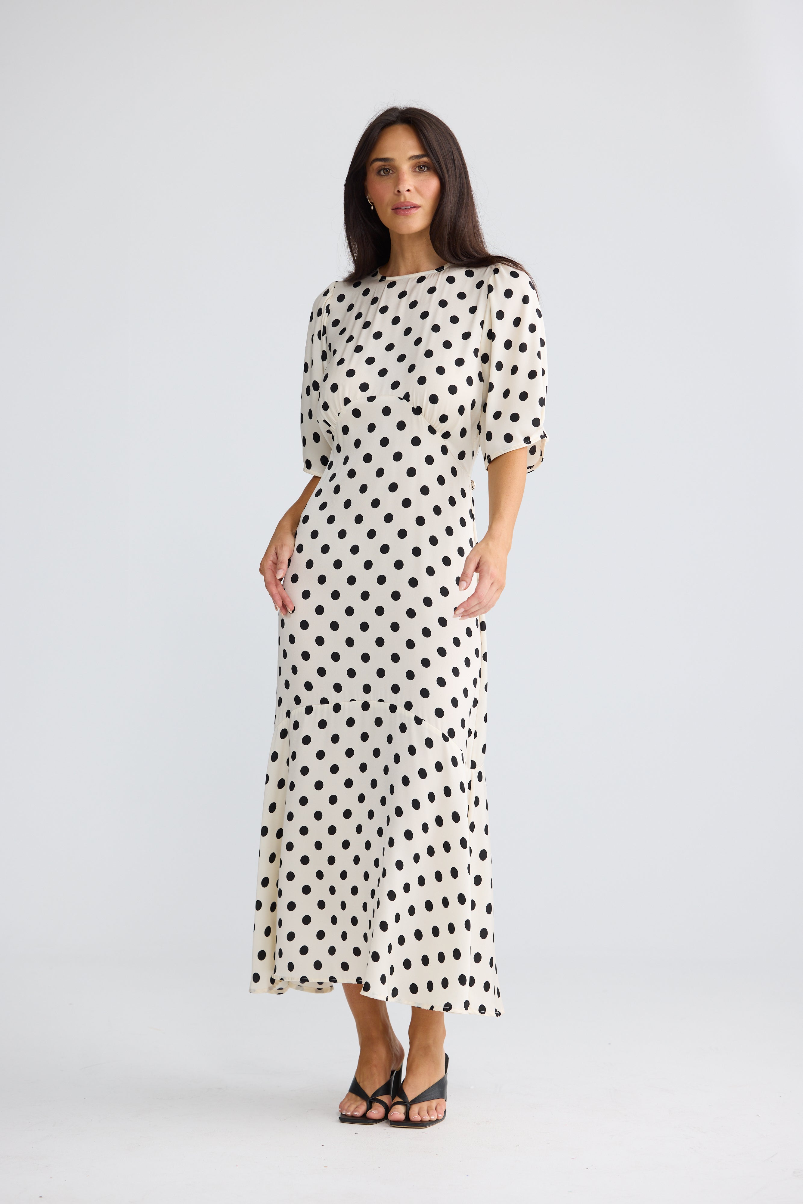 Image of A woman modeling a Brave & True midi-length cream dress with black polka dots, featuring elbow-length puff sleeves and a boat neckline, paired with black strappy heels against a light gray studio background.