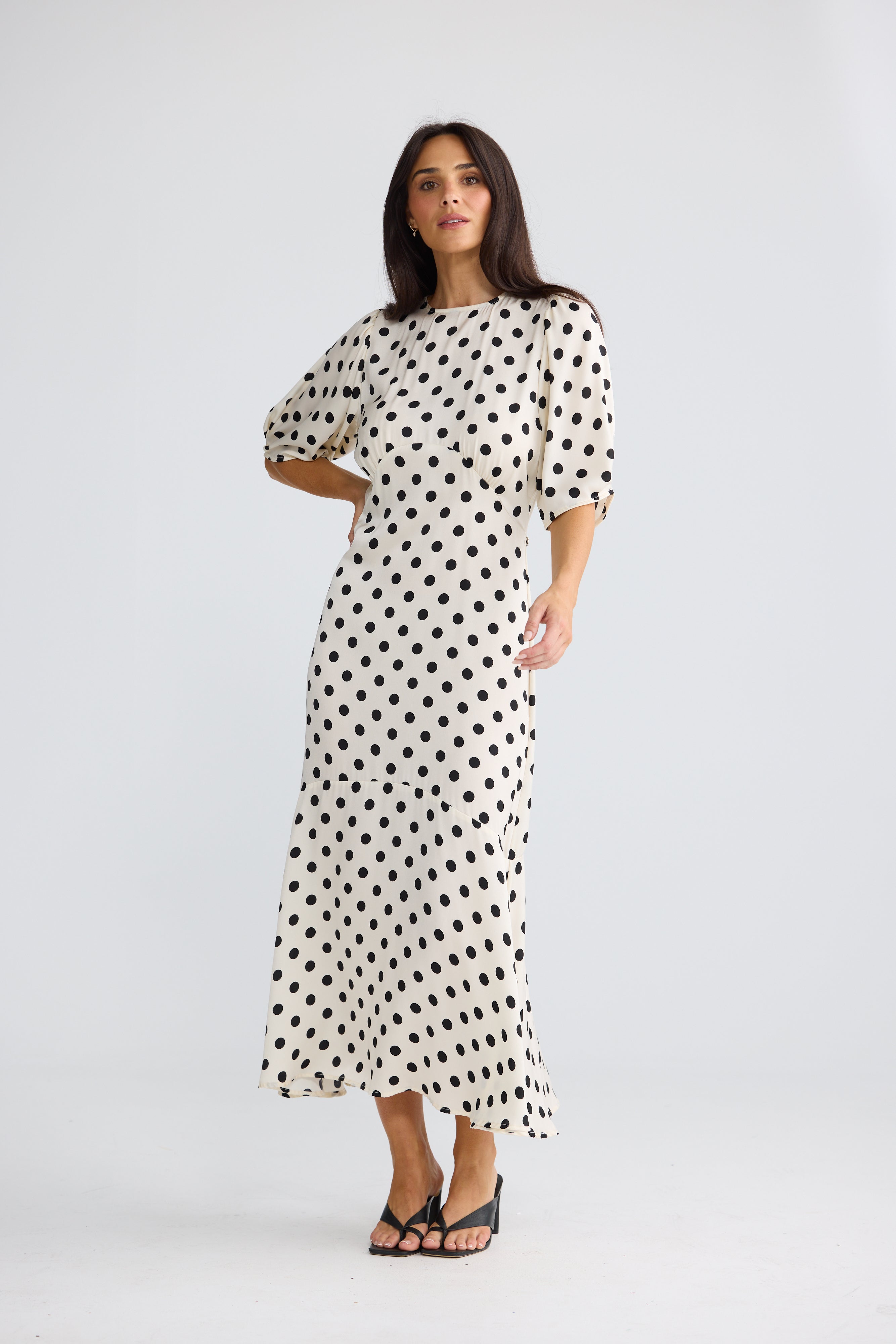 Image of A woman modeling a Brave & True midi-length white dress with a black polka dot pattern, featuring short puff sleeves and a boat neckline, photographed against a light gray studio background.