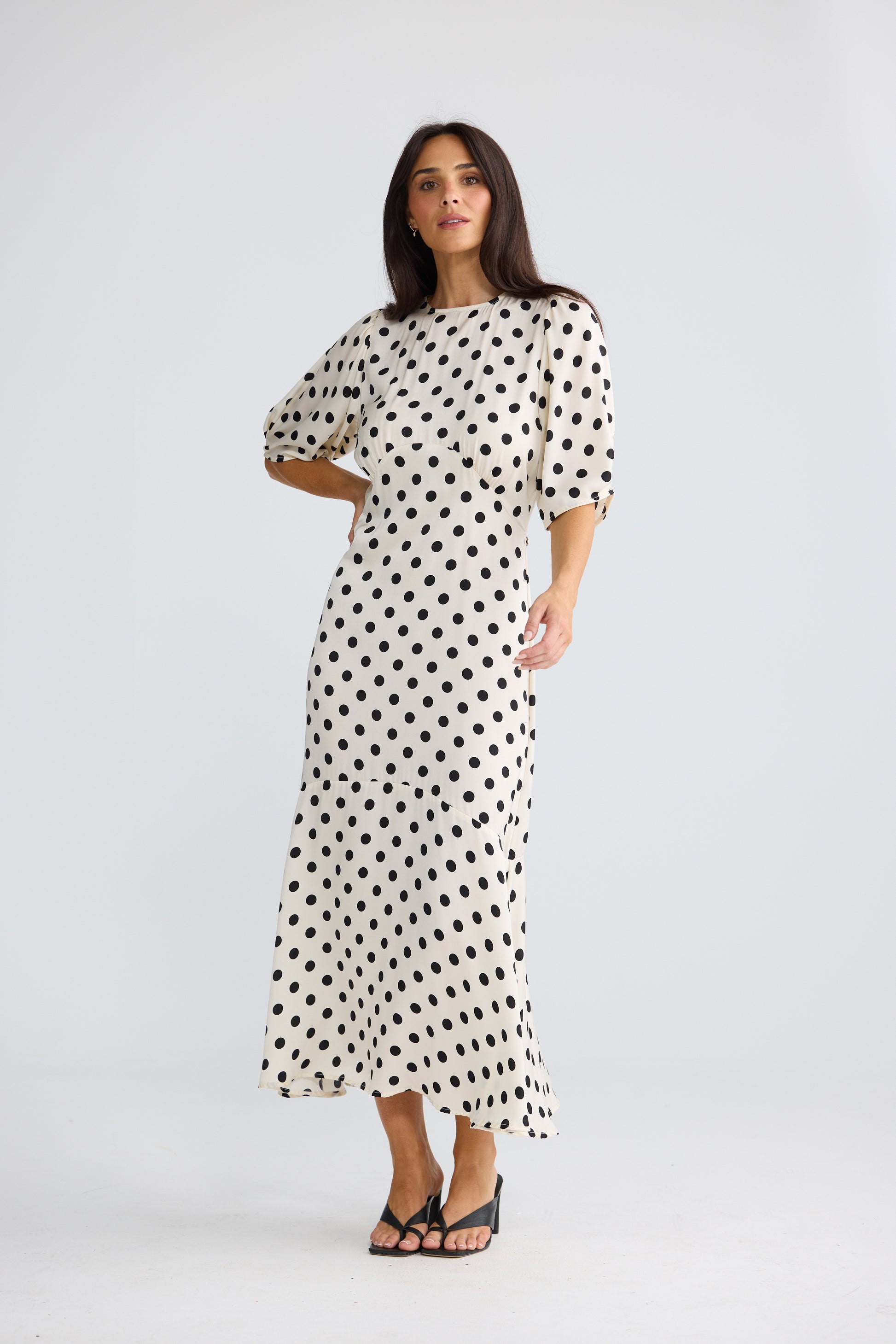 A woman modeling a Brave & True midi-length white dress with a black polka dot pattern, featuring short puff sleeves and a boat neckline, photographed against a light gray studio background.