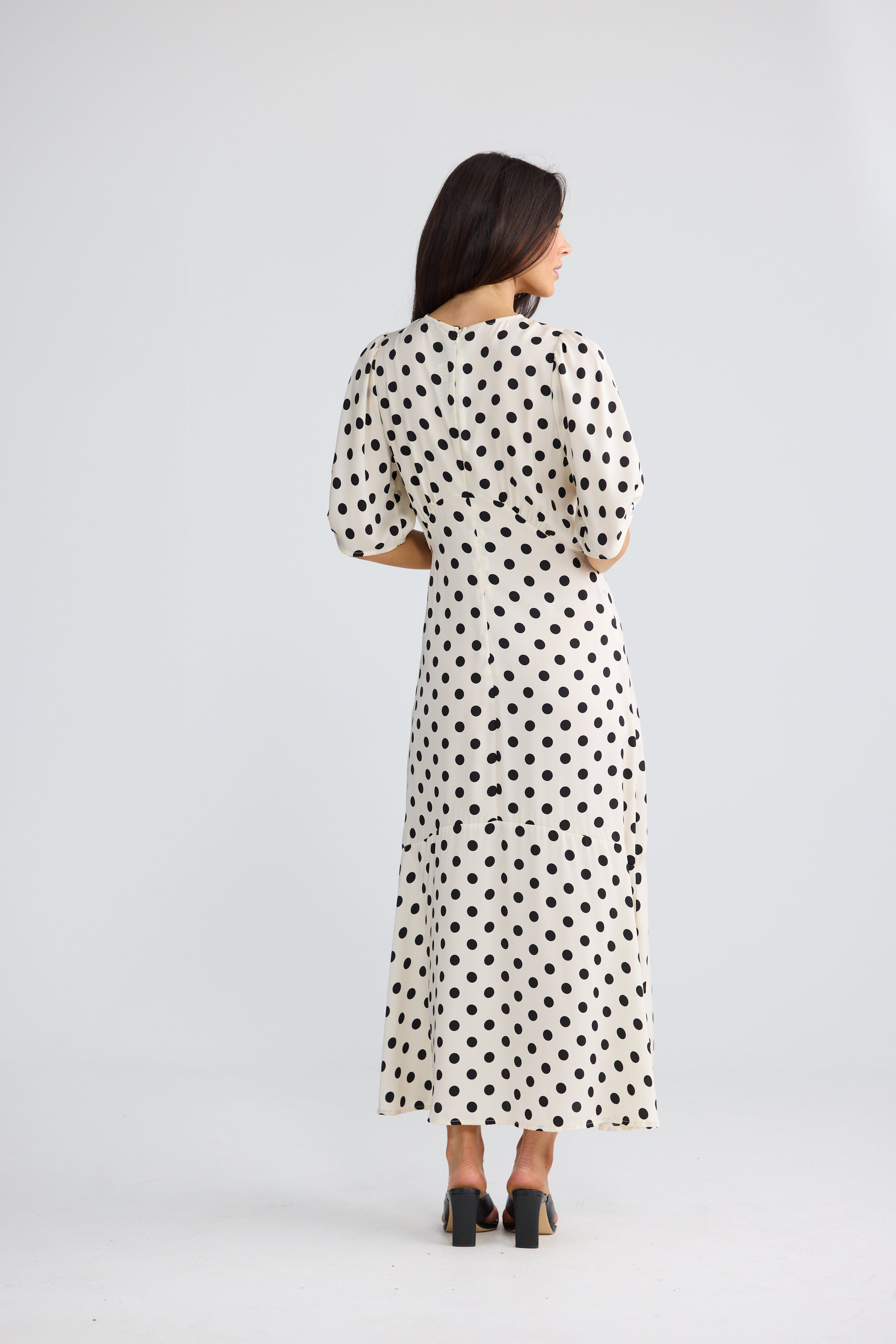 Image of A woman viewed from the back wearing a Brave & True cream-colored midi dress with black polka dots, three-quarter sleeves, and black high heels, set against a plain light gray studio background.