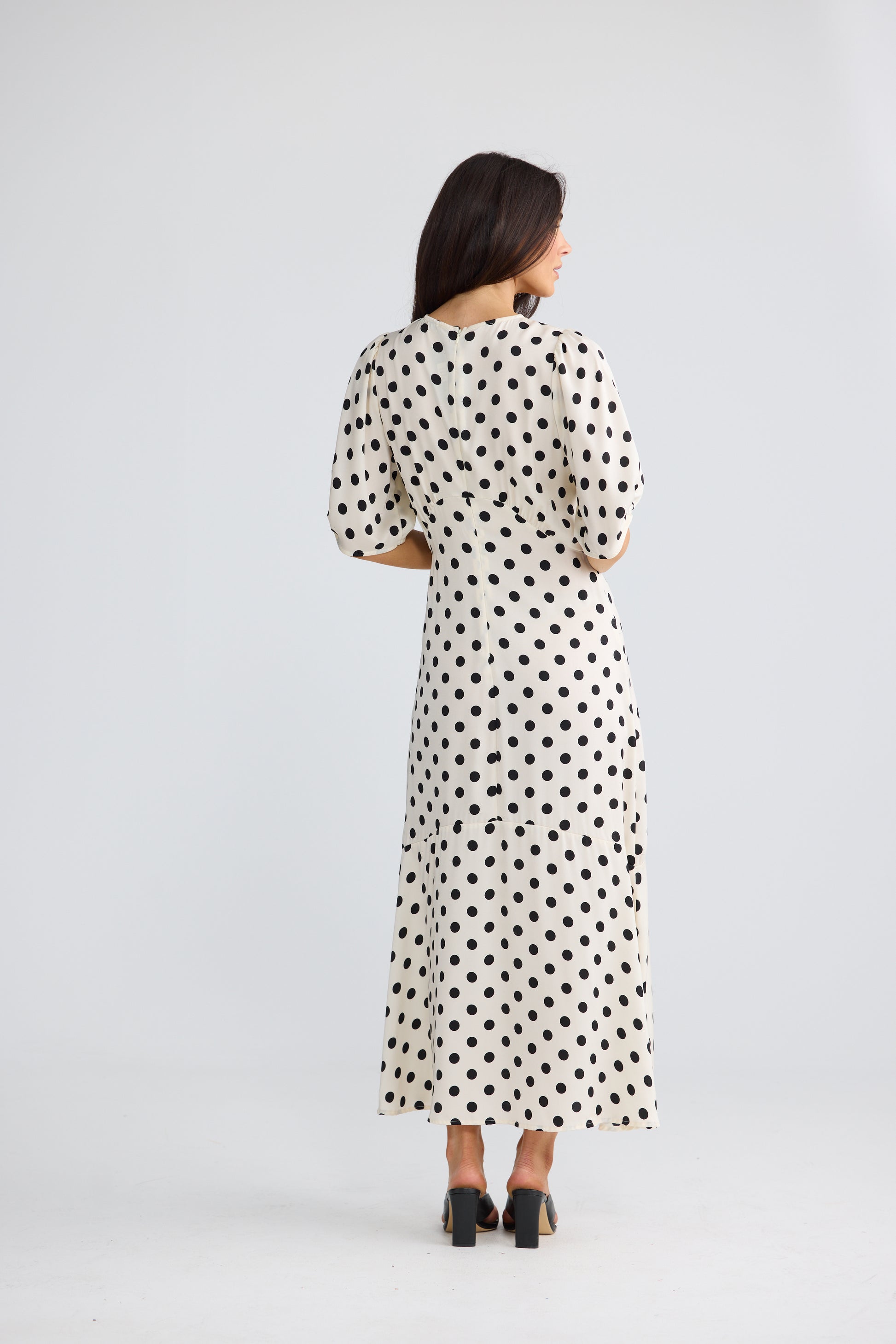A woman viewed from the back wearing a Brave & True cream-colored midi dress with black polka dots, three-quarter sleeves, and black high heels, set against a plain light gray studio background.