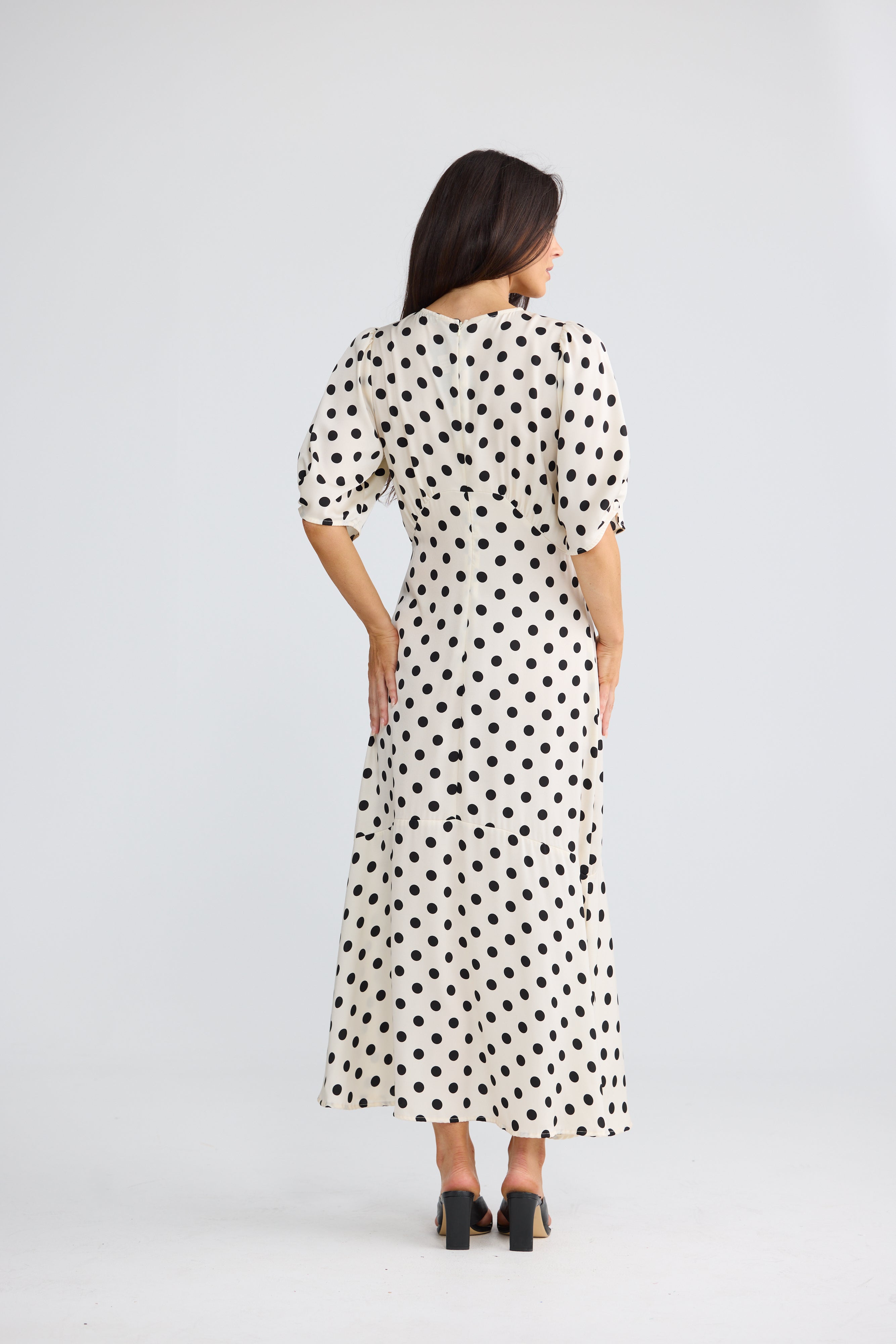 Image of A woman viewed from the back wearing a Brave & True midi dress with puff sleeves and an allover black polka dot pattern on a white base, paired with black heels, against a light gray studio background.