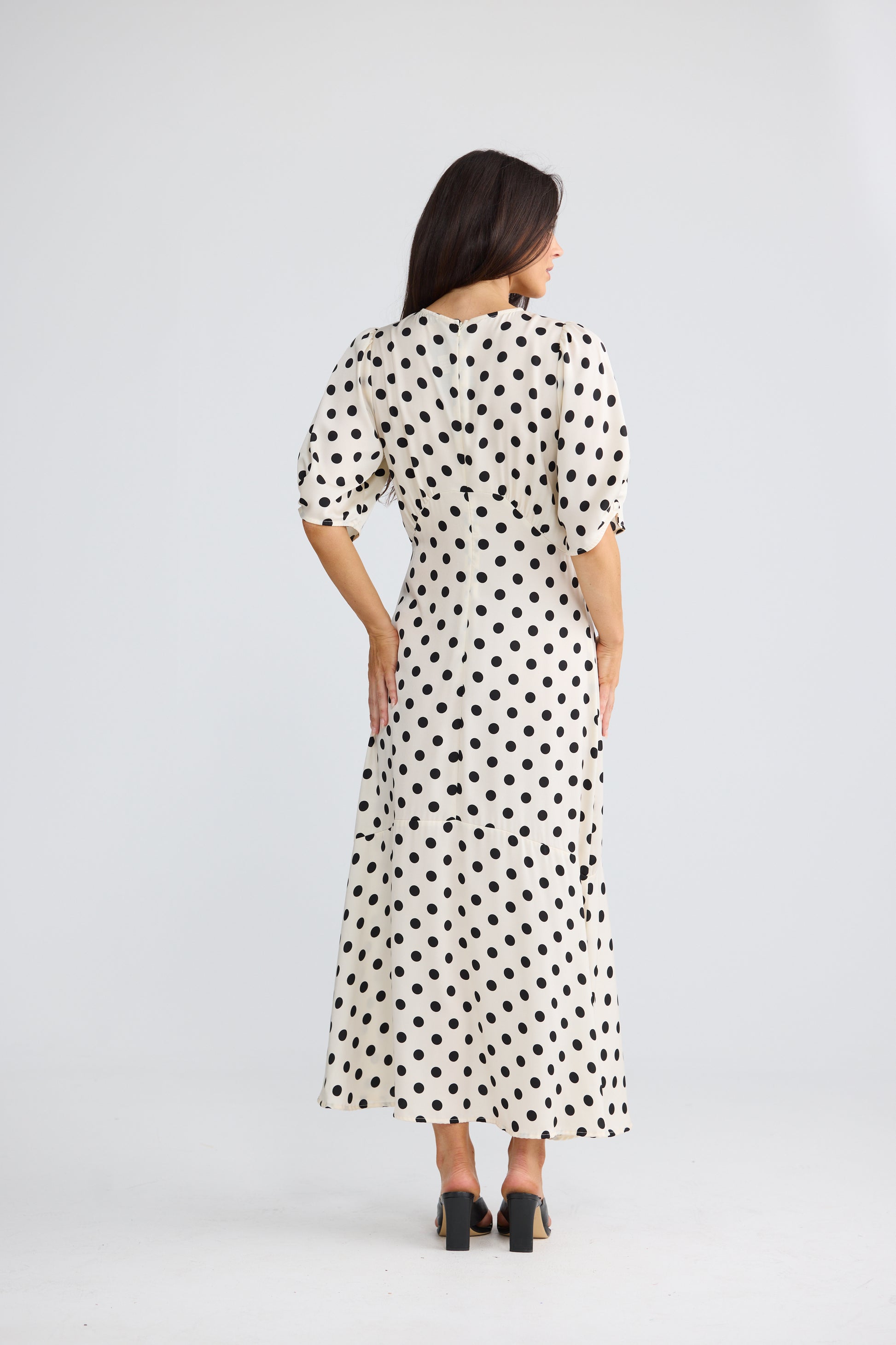 A woman viewed from the back wearing a Brave & True midi dress with puff sleeves and an allover black polka dot pattern on a white base, paired with black heels, against a light gray studio background.
