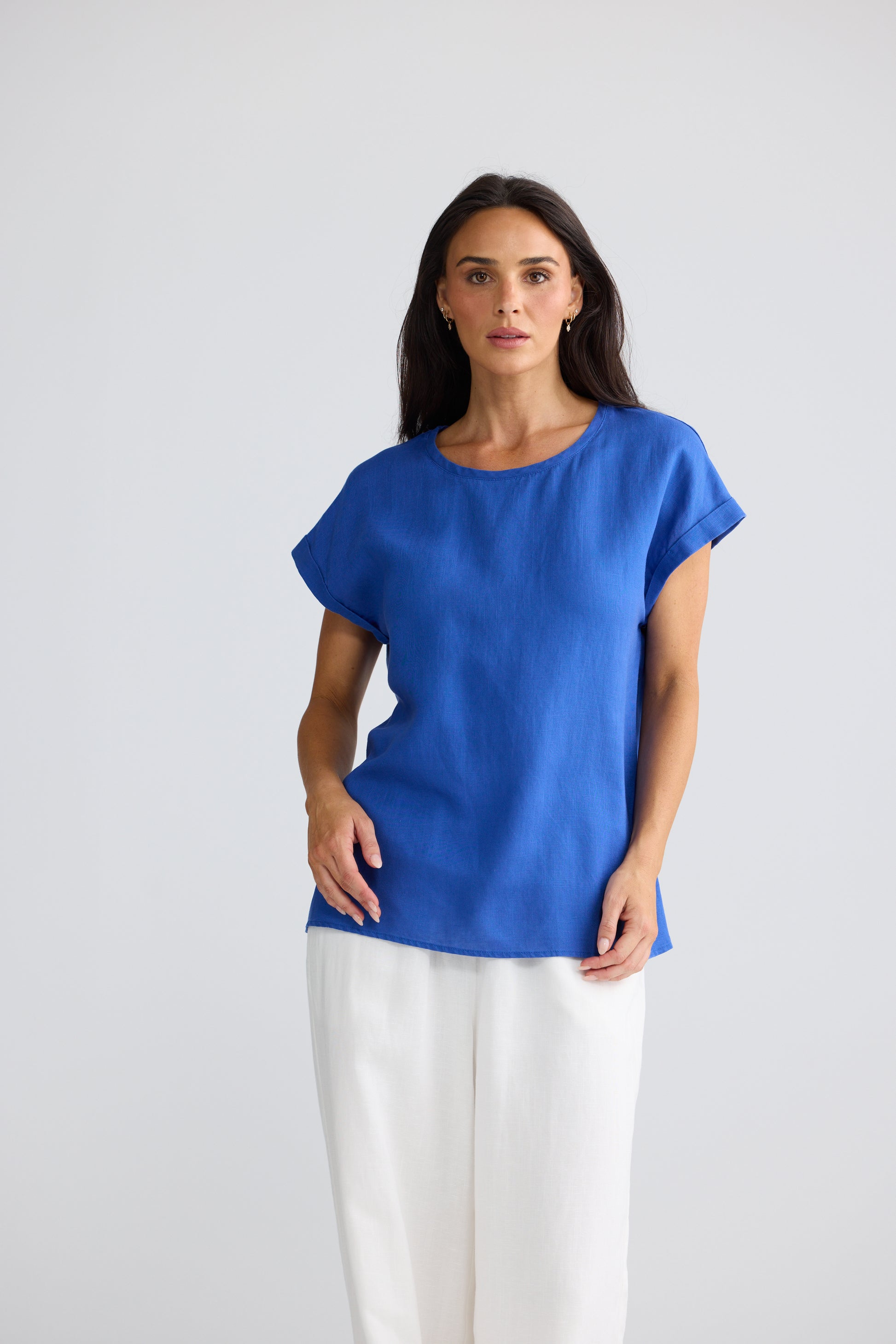 A woman modeling a Brave & True short-sleeve, crew-neck top in a vibrant royal blue, paired with white wide-leg trousers, against a plain light gray studio background.