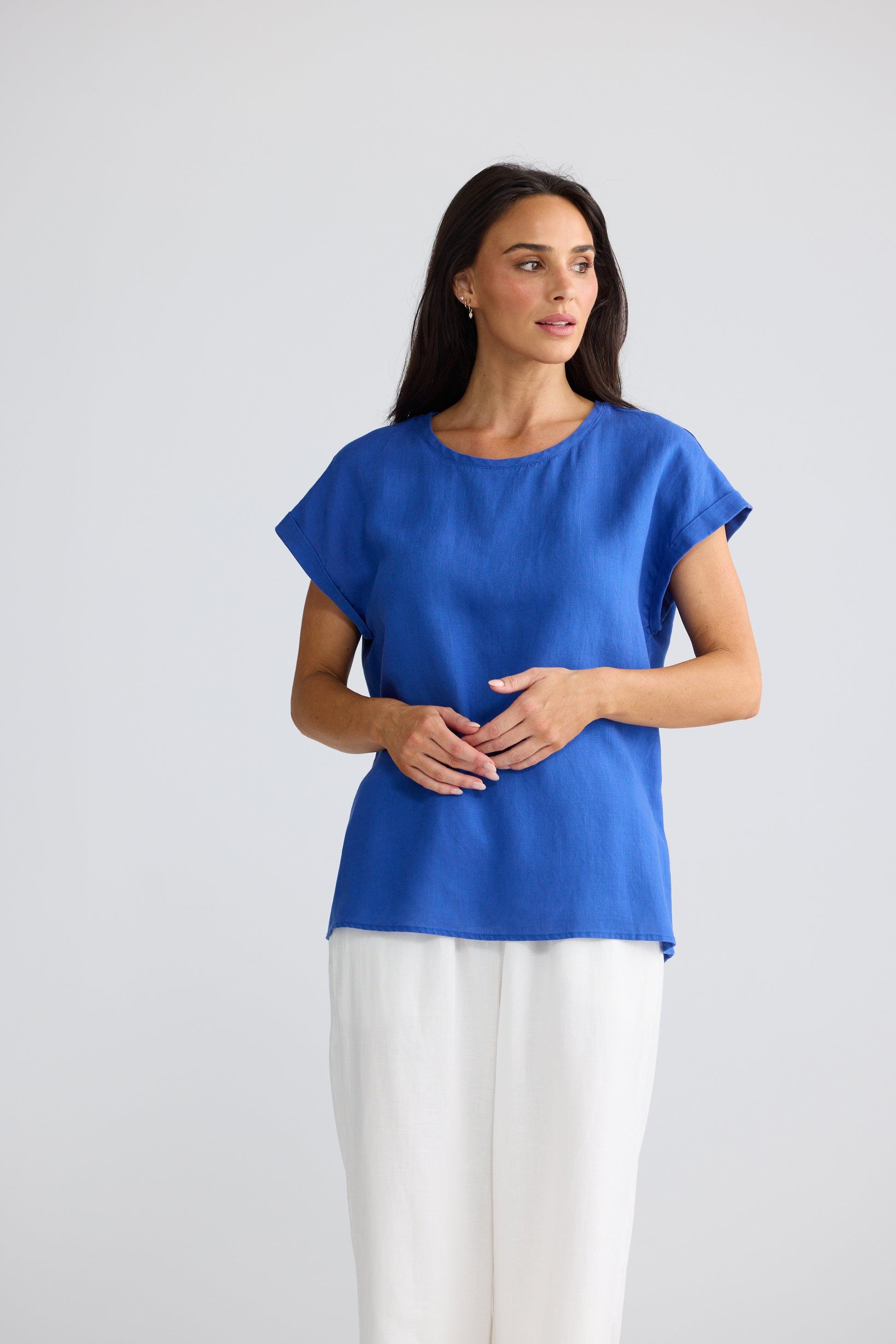 A woman modeling a Brave & True bright blue short-sleeve top with white pants, standing against a light gray studio background.