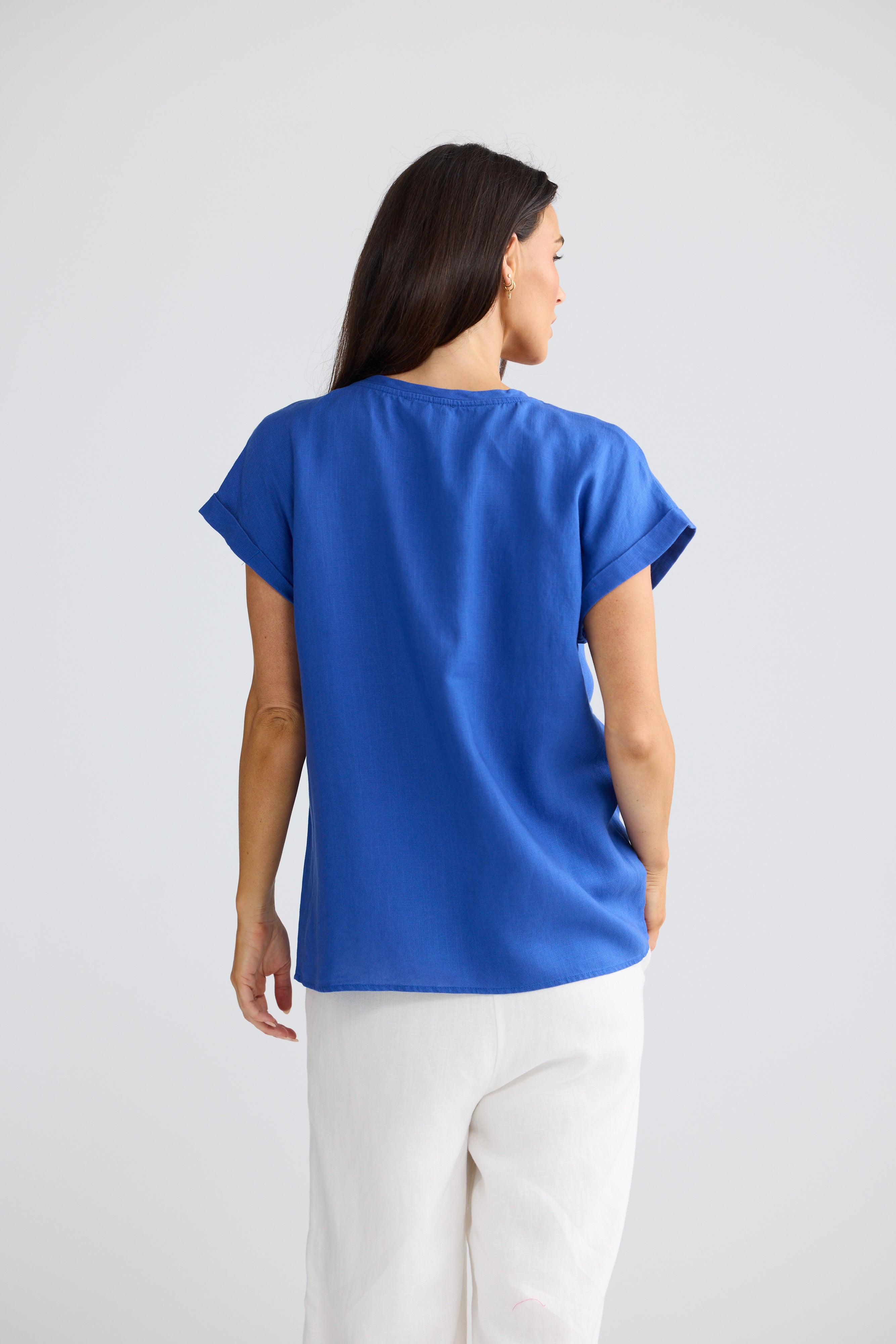 Image of A woman shown from the back wearing a Brave & True bright royal blue short-sleeve top and white pants against a light gray studio background.