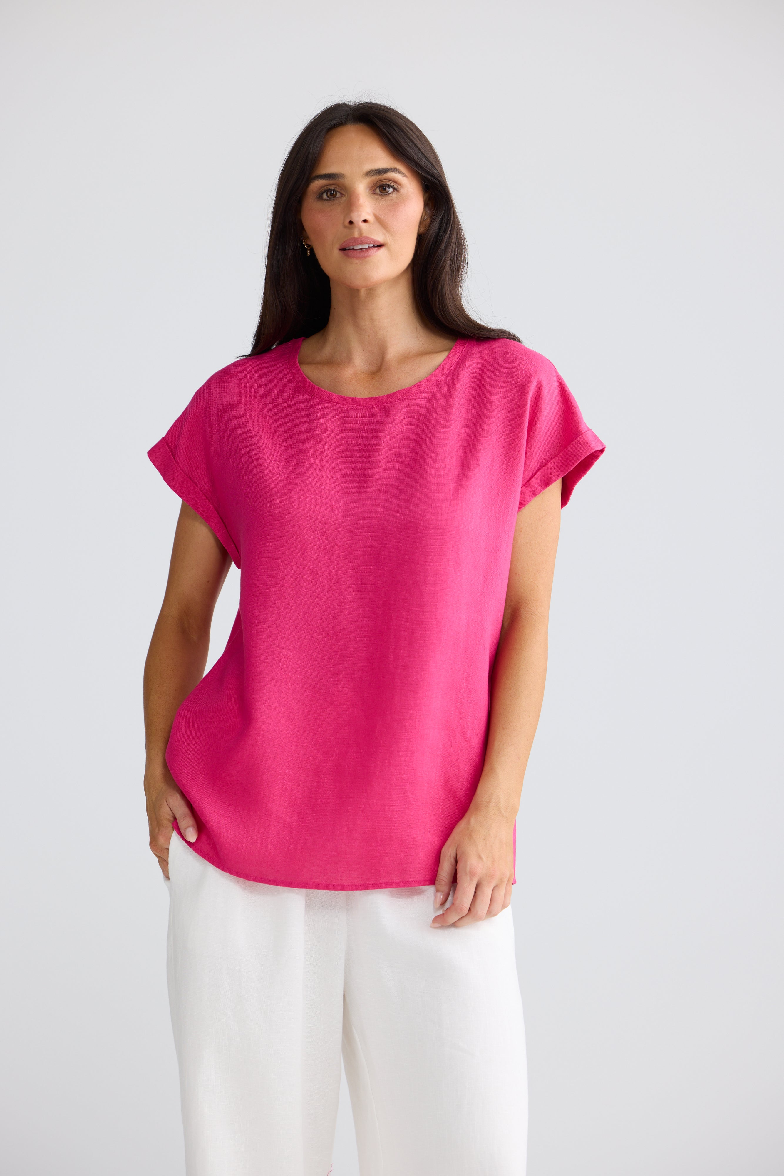 Image of A woman modeling a Brave & True bright pink short-sleeve top with a relaxed, oversized fit and white trousers, standing against a light gray studio background.