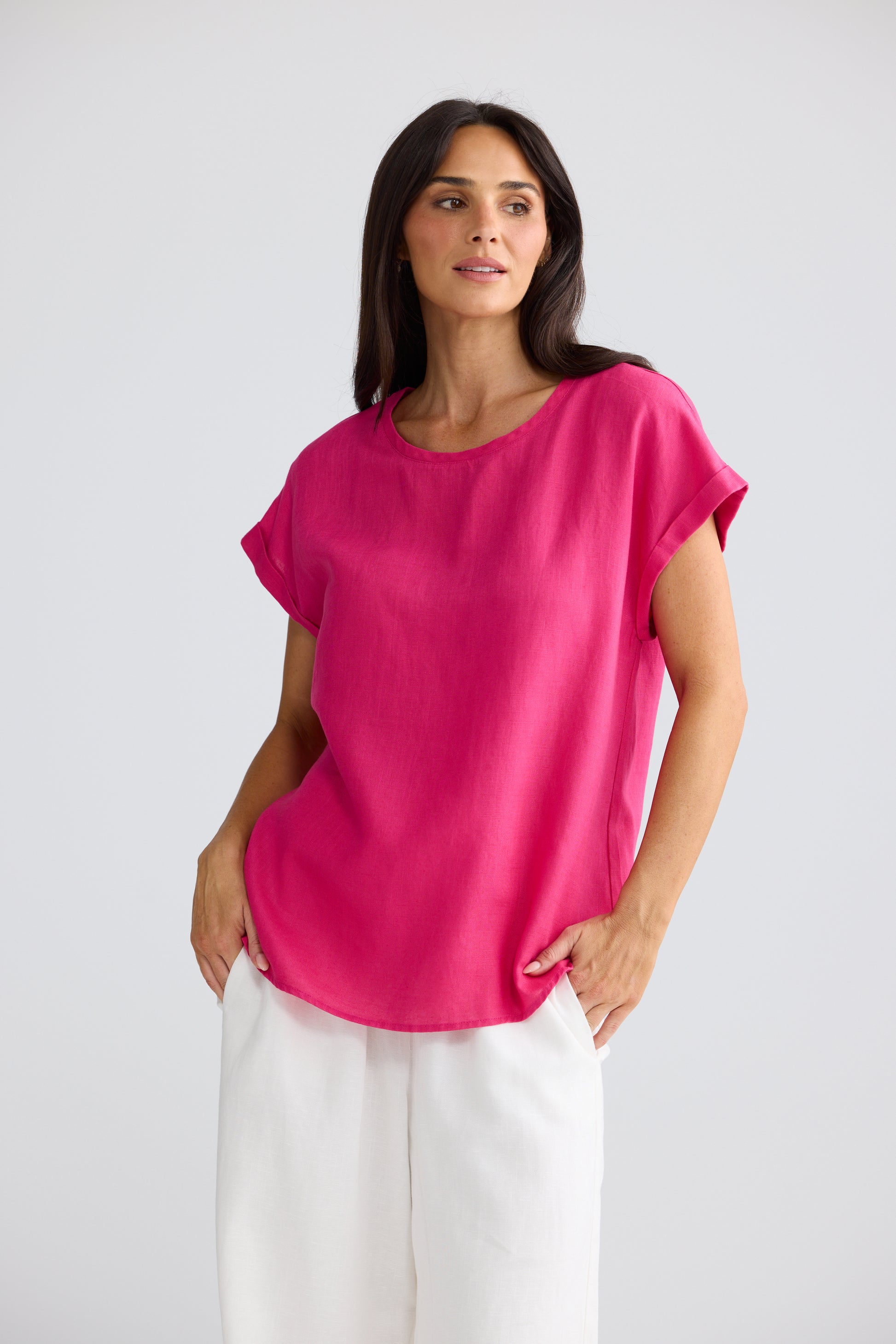 A woman modeling a Brave & True bright pink short-sleeve top with white pants, standing against a light gray studio background.
