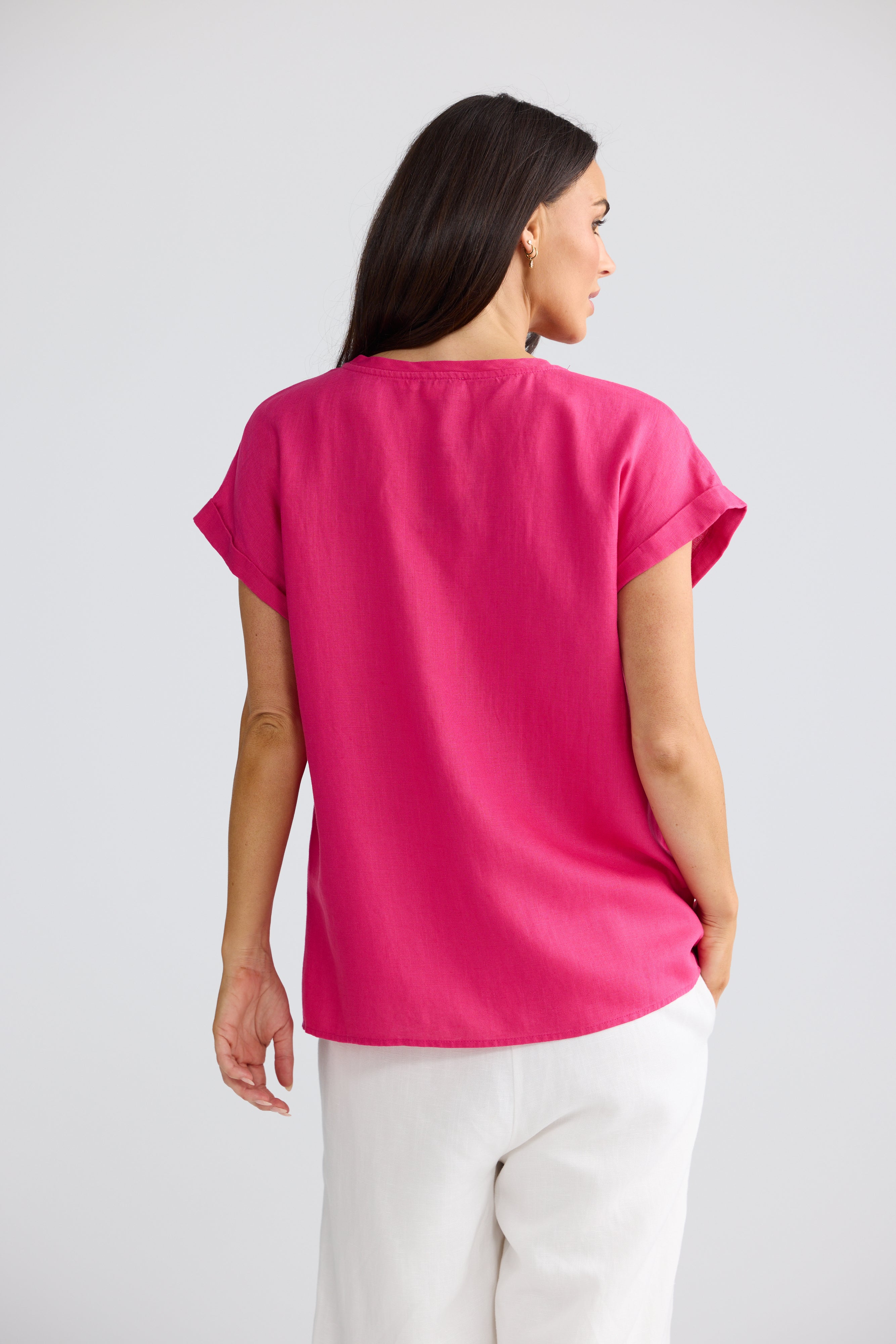Image of A woman shown from the back wearing a Brave & True bright pink short-sleeve top and white pants against a light gray studio background. 
