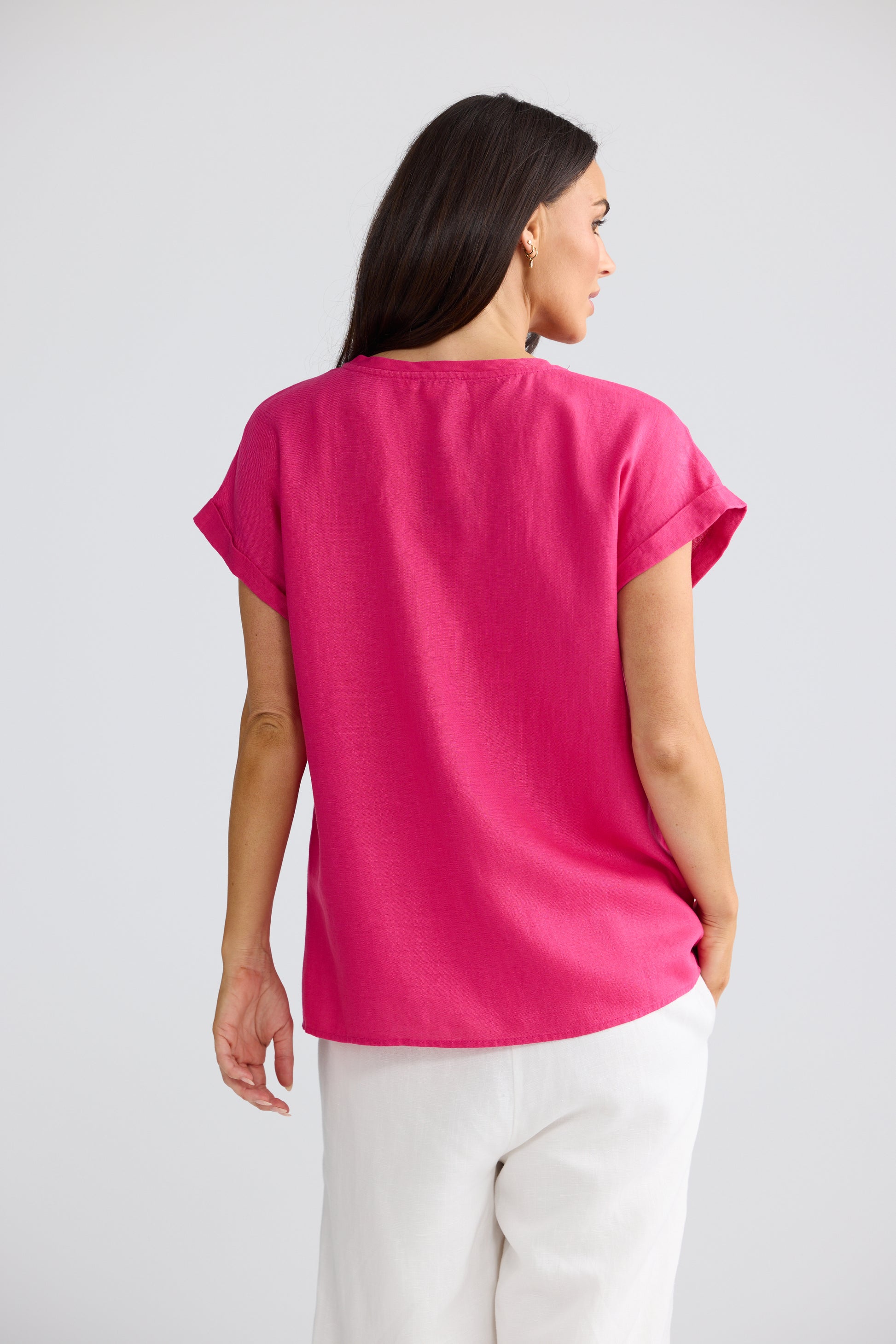 A woman shown from the back wearing a Brave & True bright pink short-sleeve top and white pants against a light gray studio background. 