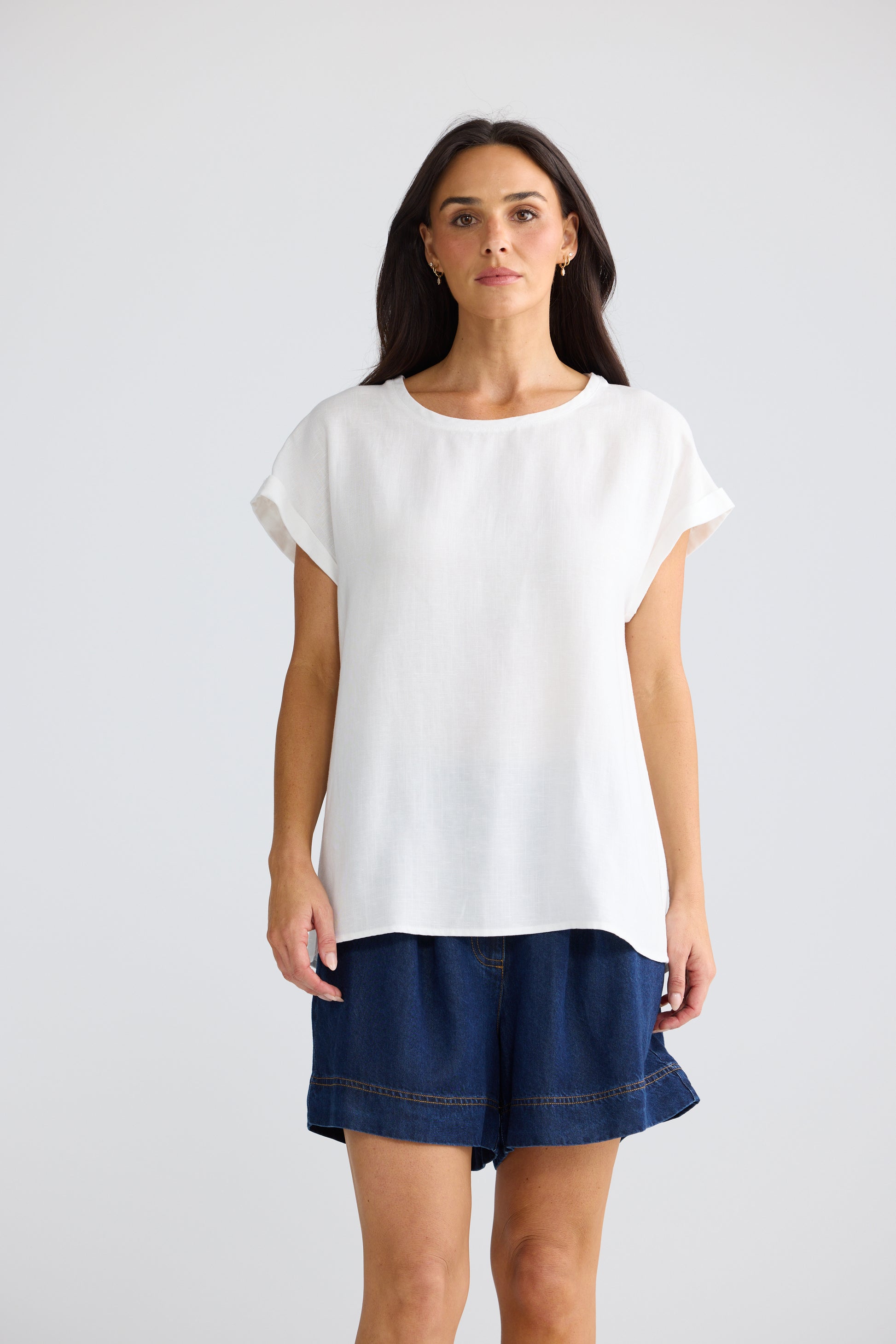 A woman modeling a Brave & True white short-sleeve crewneck top paired with dark blue denim shorts, standing against a light gray studio background.
