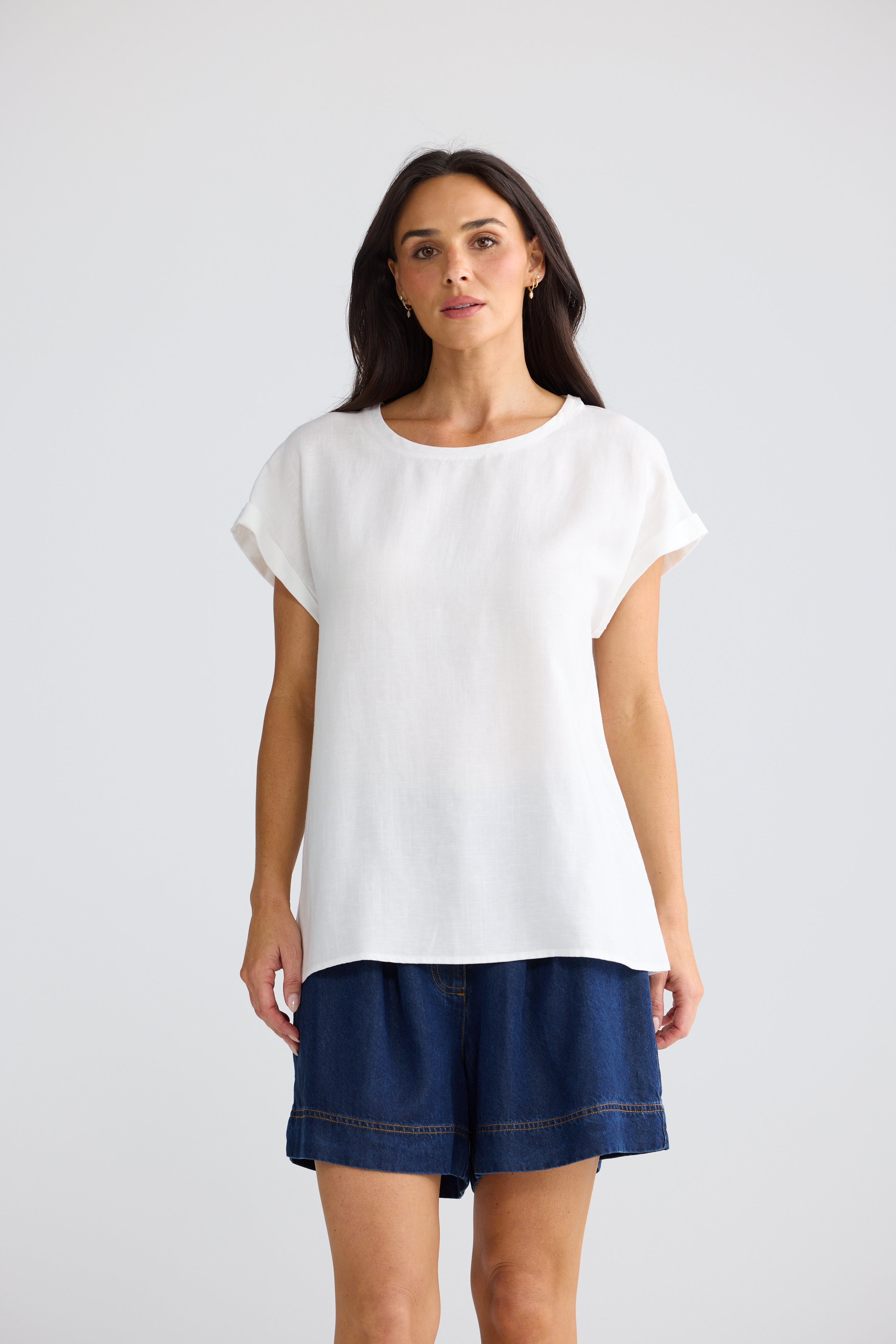 Image of A woman modeling a Brave & True white short-sleeve top with rolled cuffs and dark blue denim shorts against a light gray studio background.
