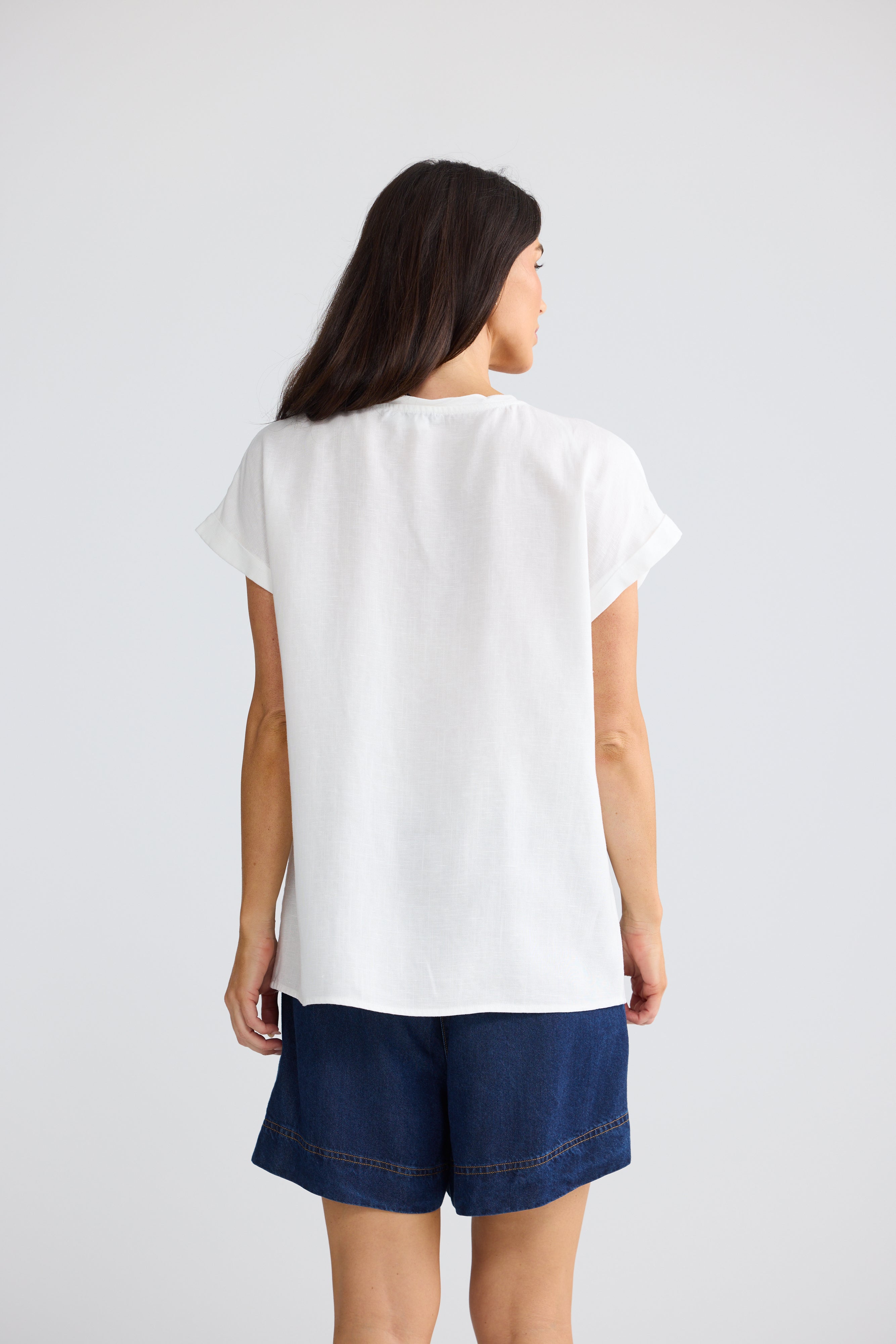 Image of A woman viewed from the back wearing a Brave & True loose white short-sleeve top and dark blue denim shorts against a light gray studio background.
