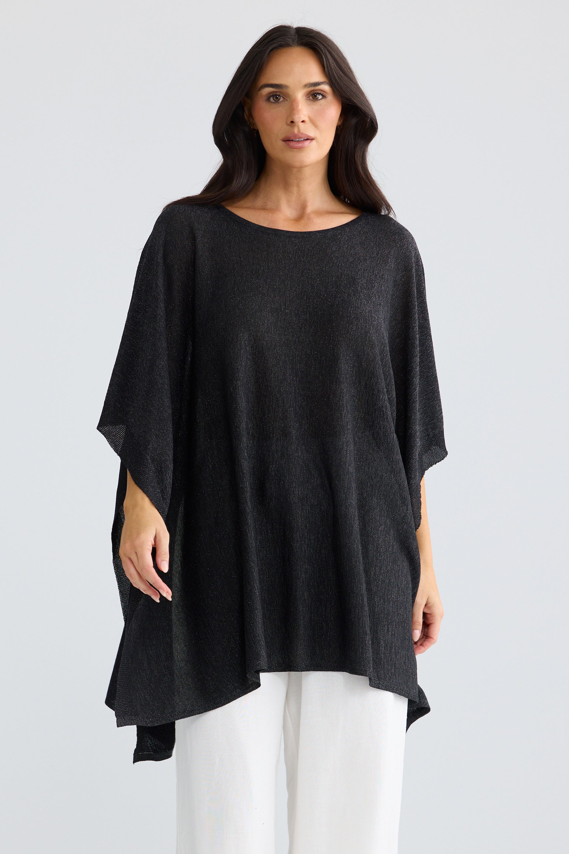 A woman modeling a Brave & True  loose, black, short-sleeved tunic top with a wide boat neckline, paired with white pants, against a light gray studio background.