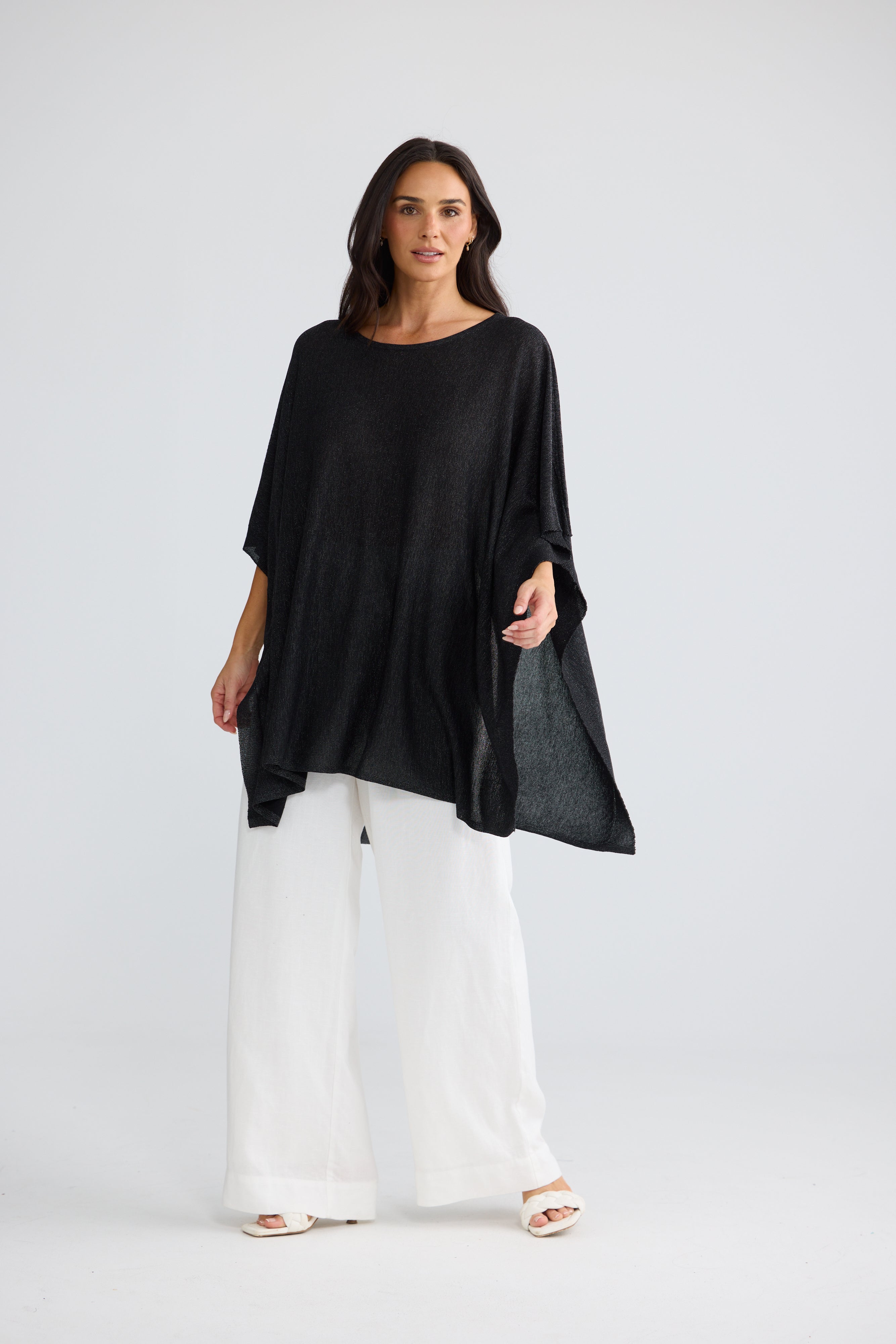 Image of A woman modeling a Brave & True loose-fitting black short-sleeve top with wide draped sleeves, paired with white wide-leg pants and white sandals, against a light gray studio background.