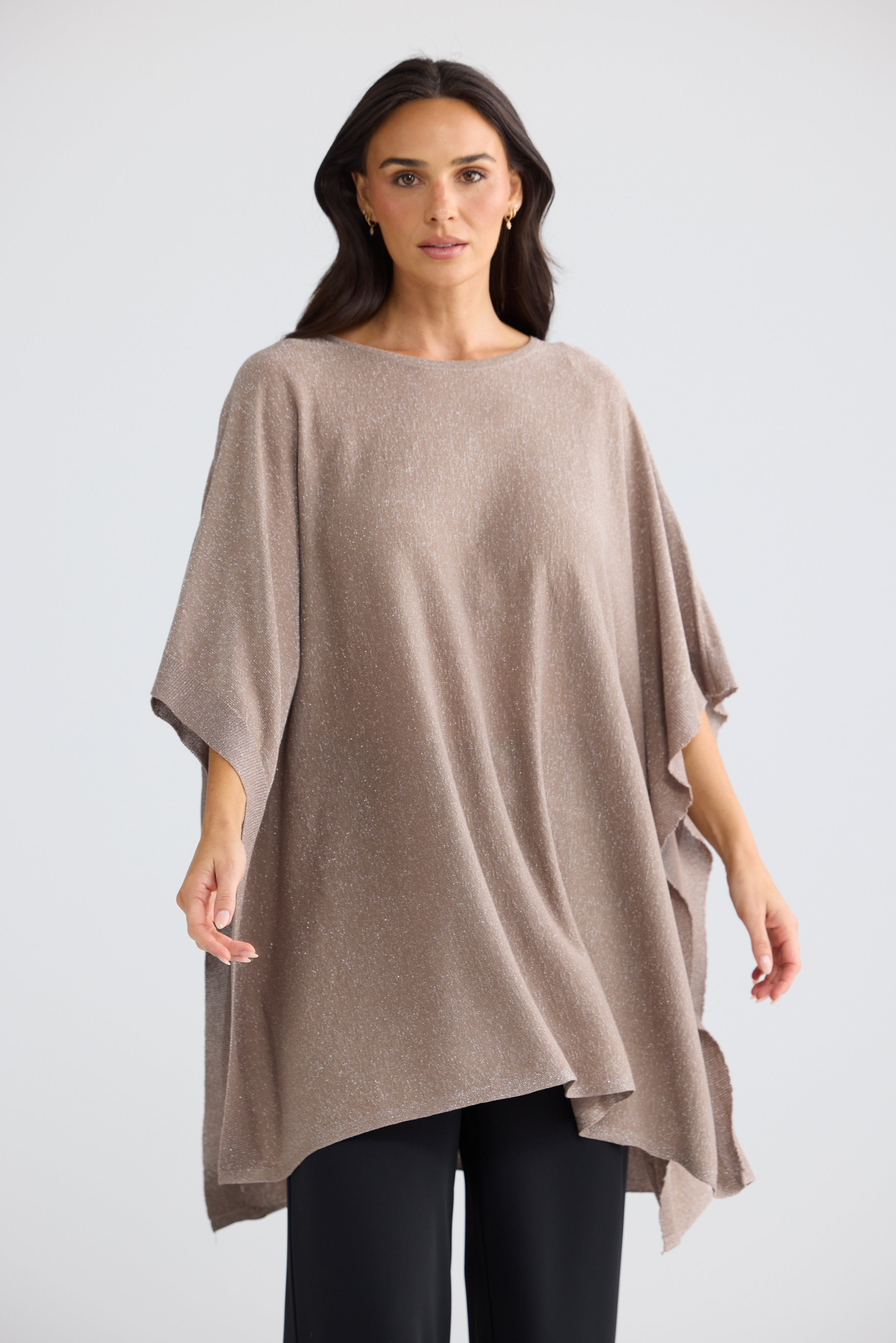 Image of A woman modeling a Brave & True loose, draped knit poncho top in a taupe-beige color, paired with black pants, against a light gray studio background.
