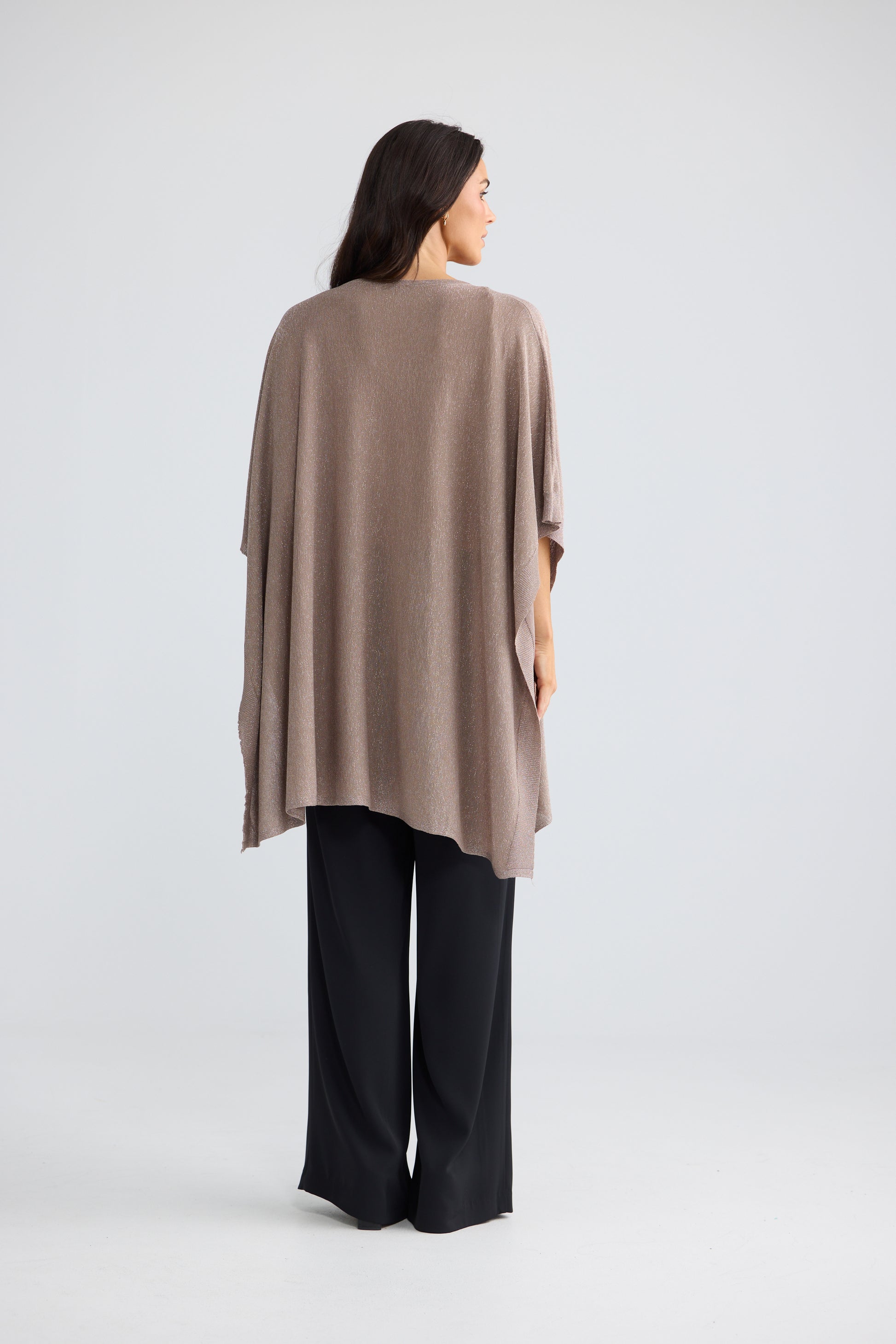 A woman shown from the back wearing a Brave & True draped, short-sleeved taupe knit poncho over black wide-leg trousers, photographed against a seamless light gray studio background.