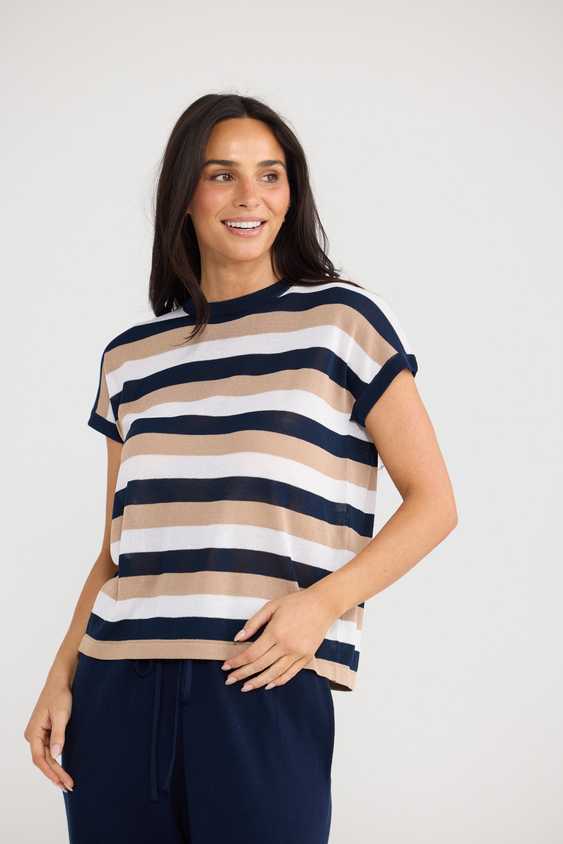 A woman wearing a Brave & True short-sleeve crewneck knit top with horizontal stripes in navy blue, beige, and white, paired with navy pants, against a light gray studio background.