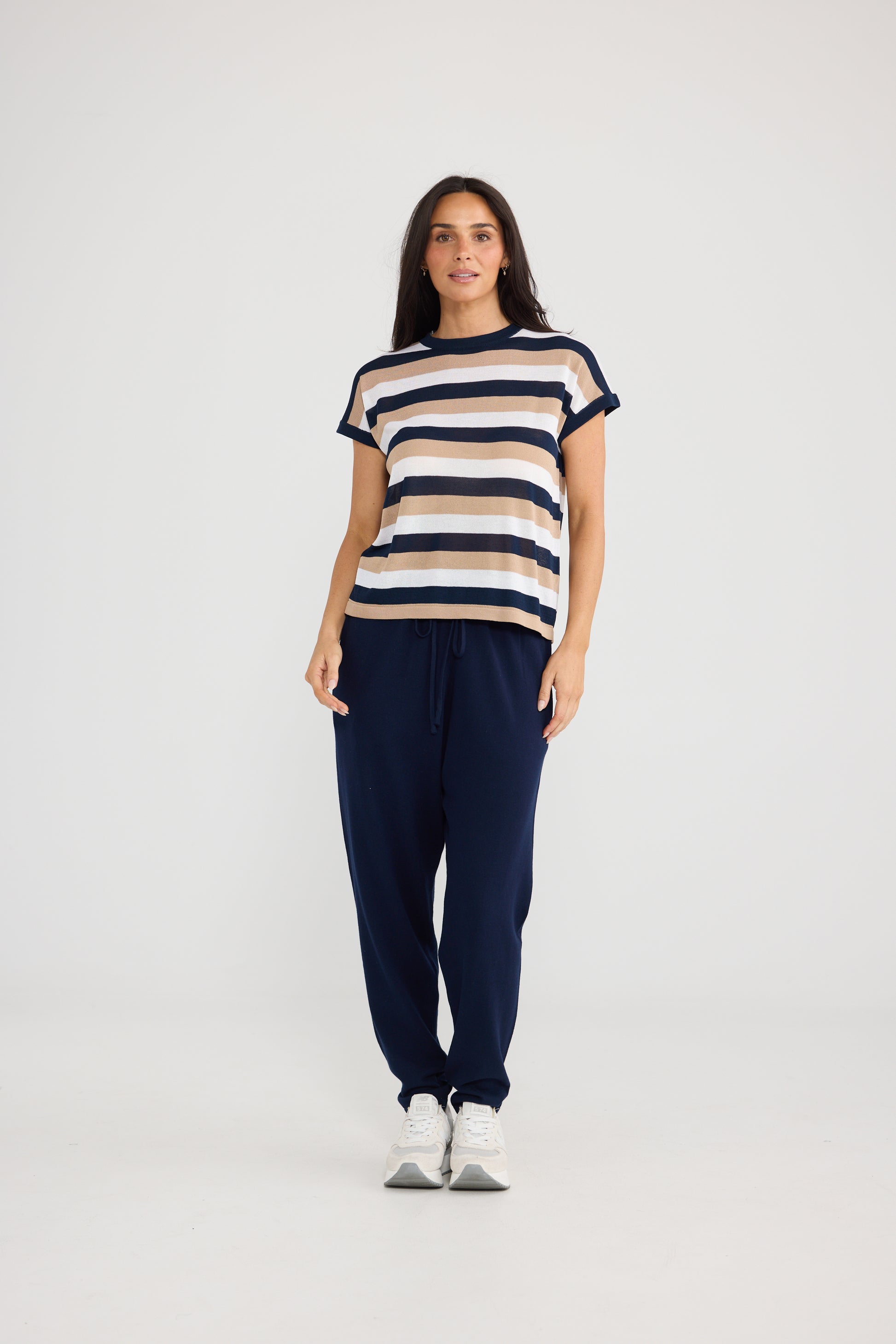 A woman modeling a Brave & True short-sleeve knit top with horizontal navy, cream, and tan stripes, paired with navy jogger pants and white sneakers, against a plain light gray studio background.