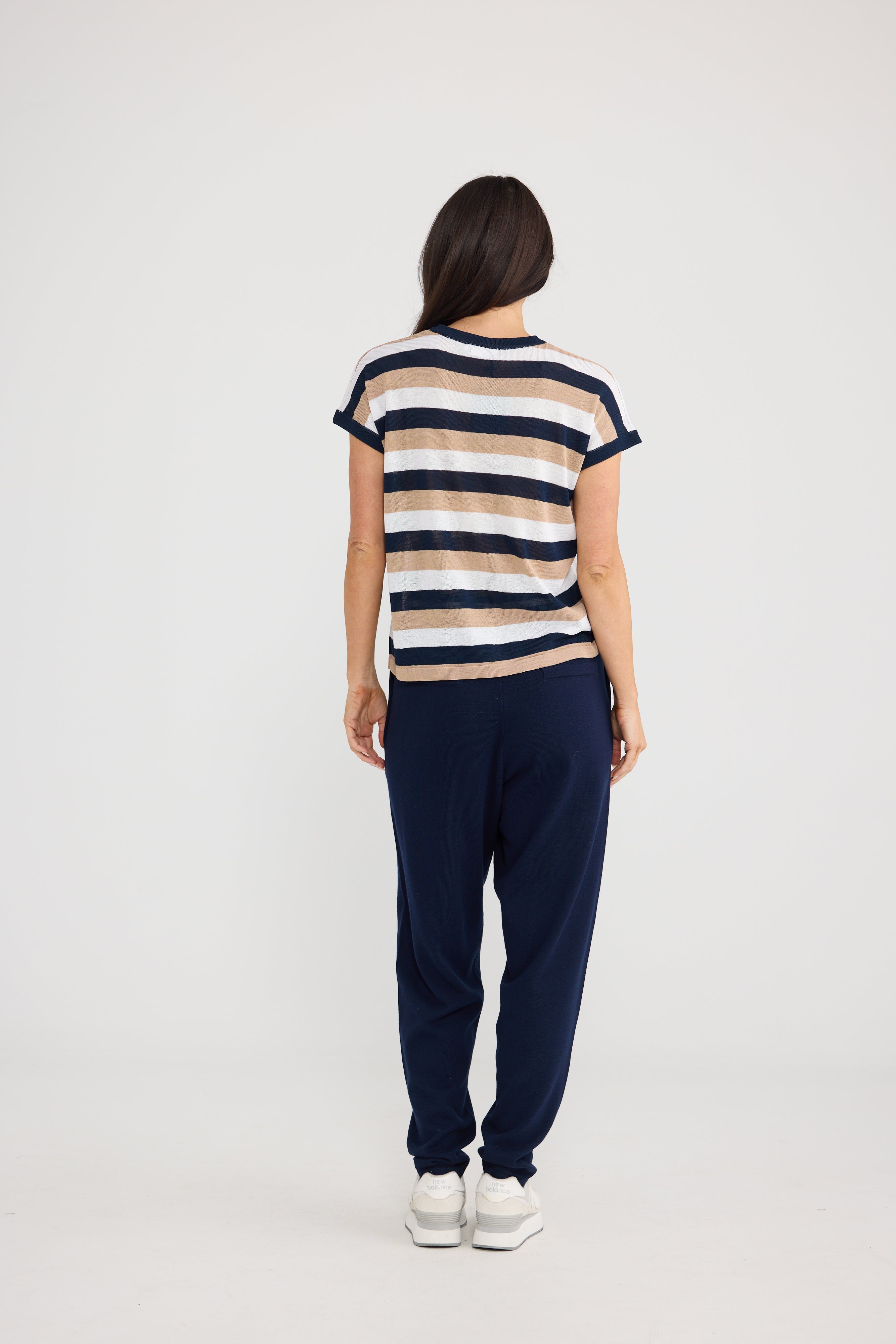 Image of A woman viewed from behind wearing a Brave & True short-sleeve striped t-shirt with navy, beige, and white horizontal stripes, paired with navy jogger pants and white sneakers, against a light gray studio background.