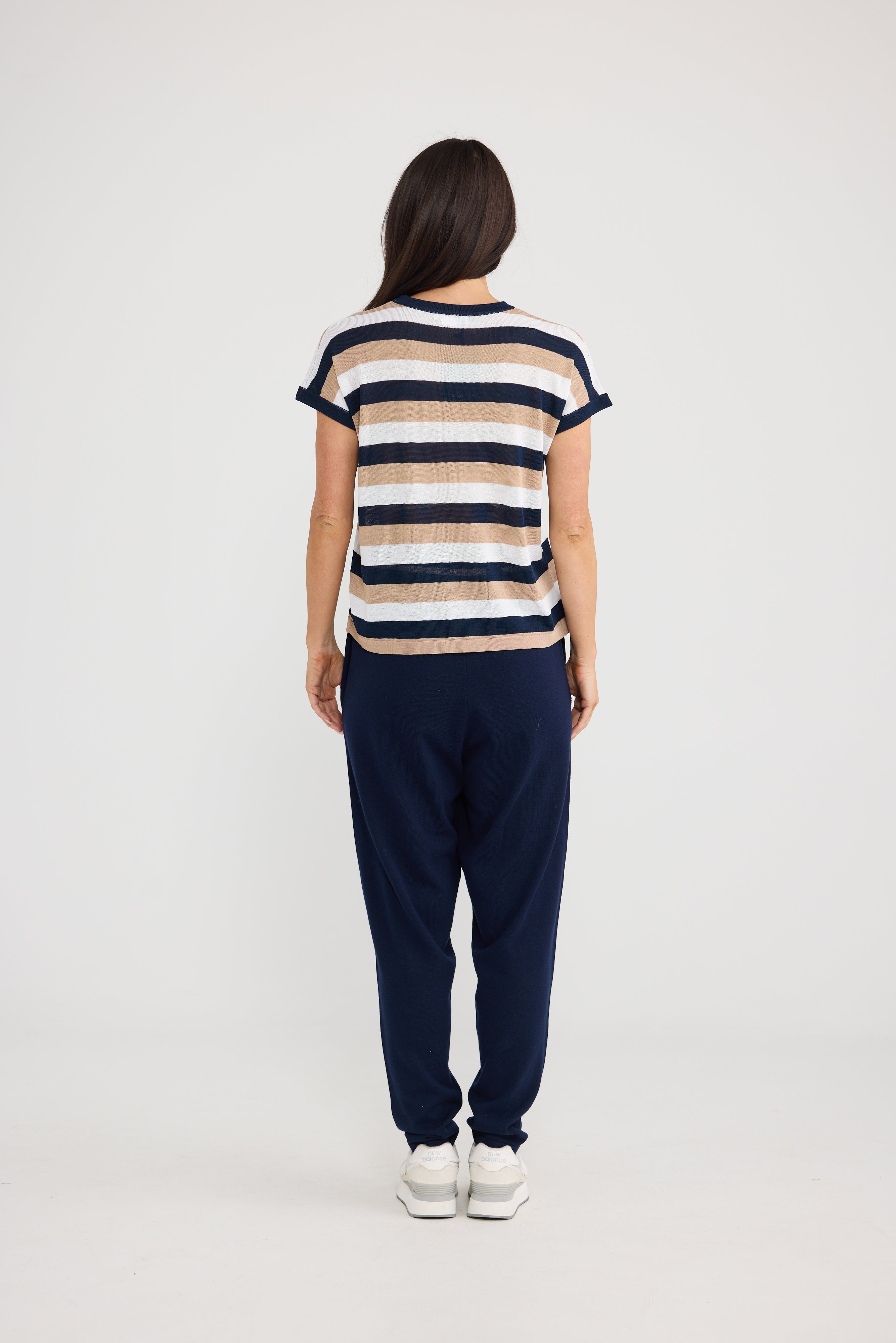 Image of A woman viewed from behind wearing a Brave & True short-sleeve striped knit top with navy, cream, and tan horizontal stripes, paired with navy jogger pants and white sneakers, against a plain light gray studio background.