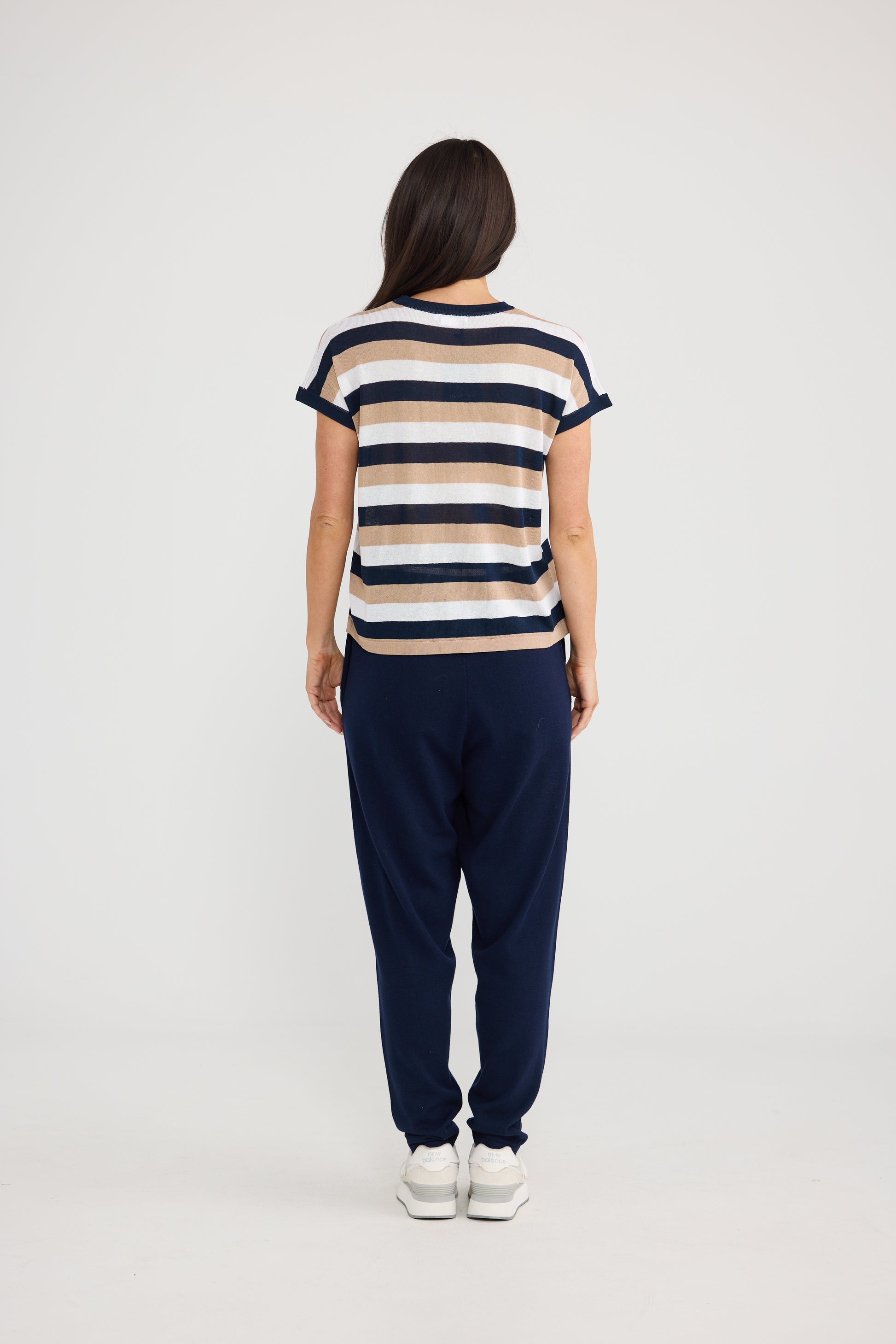A woman viewed from behind wearing a Brave & True short-sleeve striped knit top with navy, cream, and tan horizontal stripes, paired with navy jogger pants and white sneakers, against a plain light gray studio background.