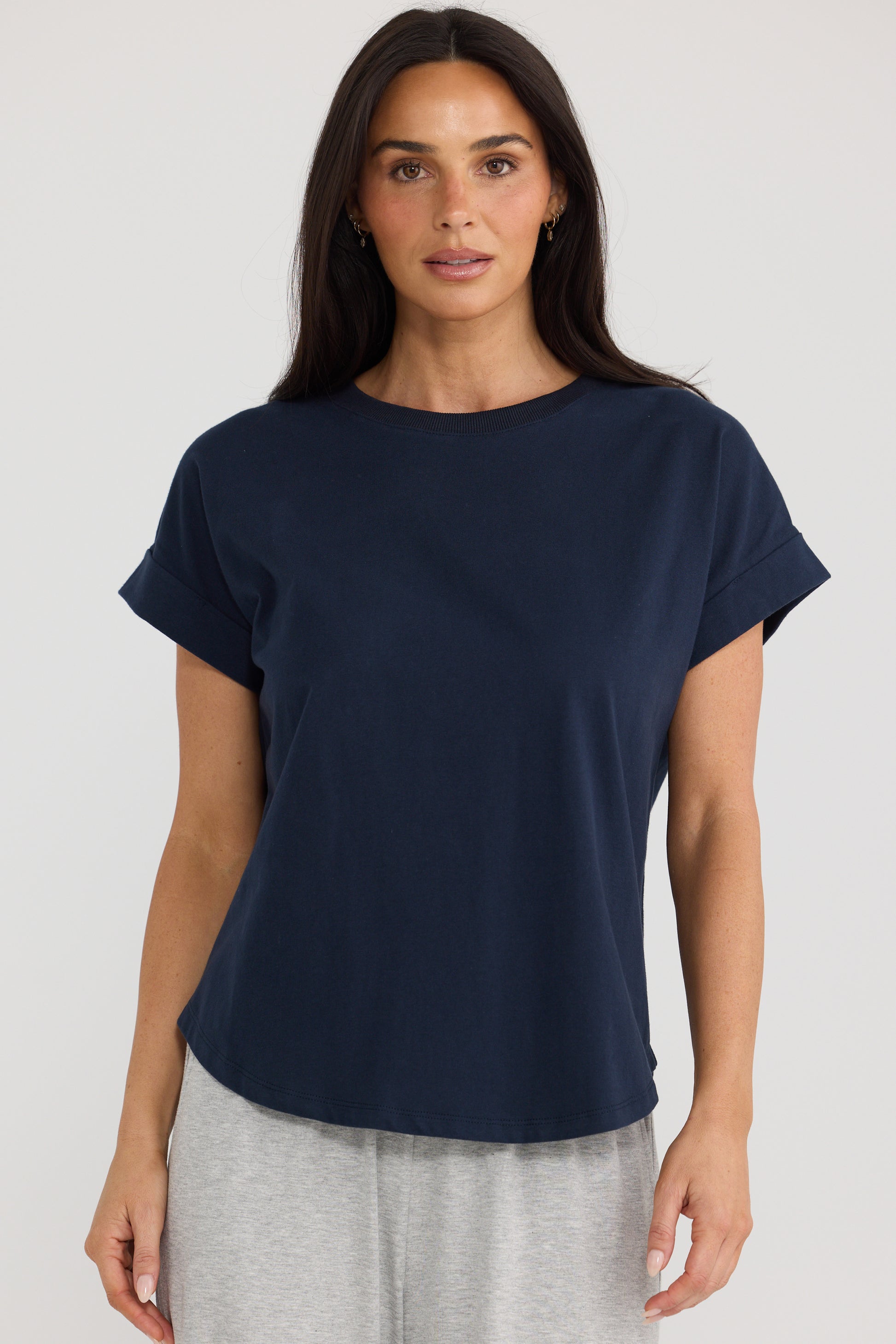 A woman wearing a Brave & True navy blue short-sleeve crewneck t-shirt and light gray sweatpants, standing against a light gray studio background.