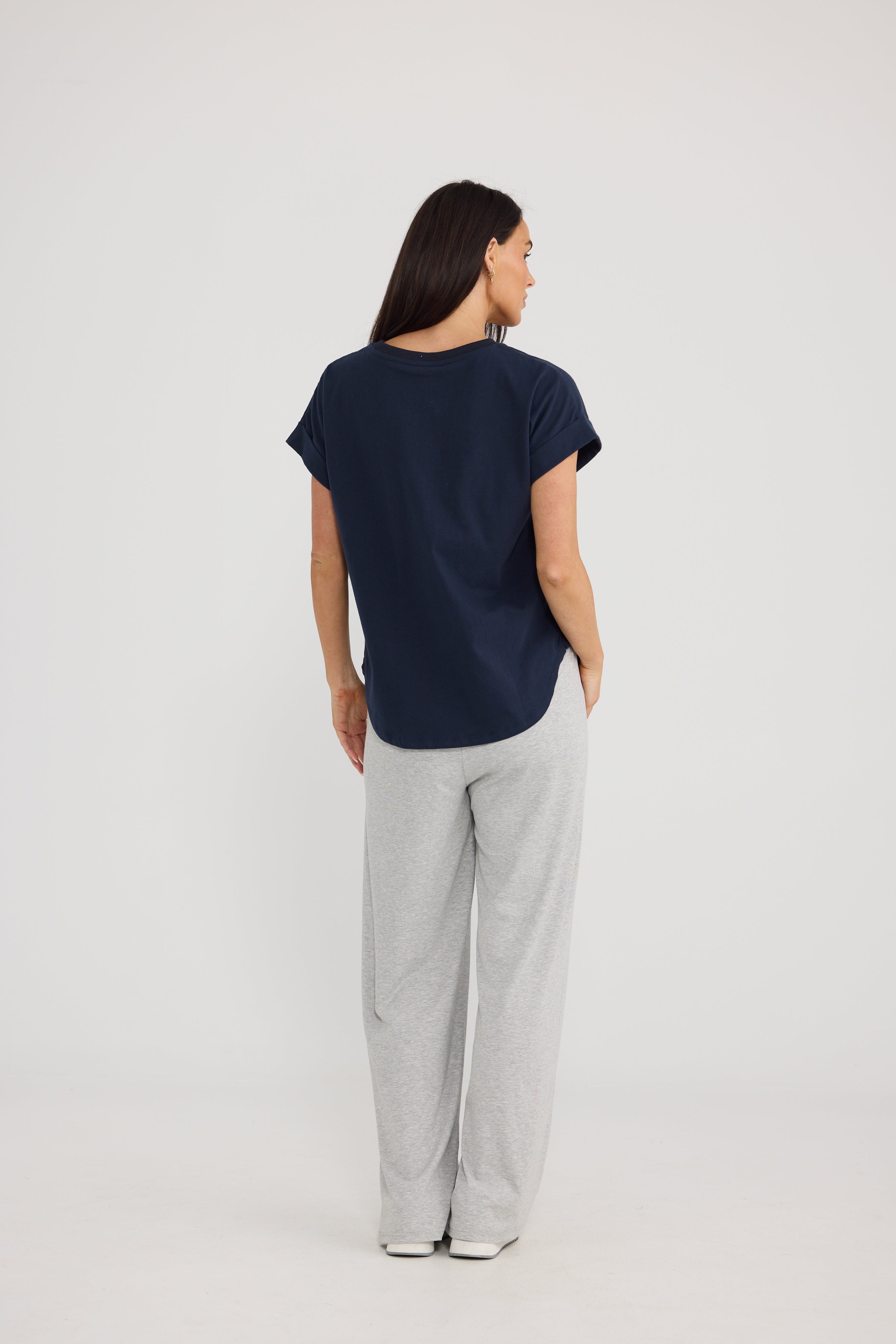 Image of A woman is shown from behind wearing a Brave & True navy blue short-sleeve top and light gray sweatpants against a plain light gray studio background.
