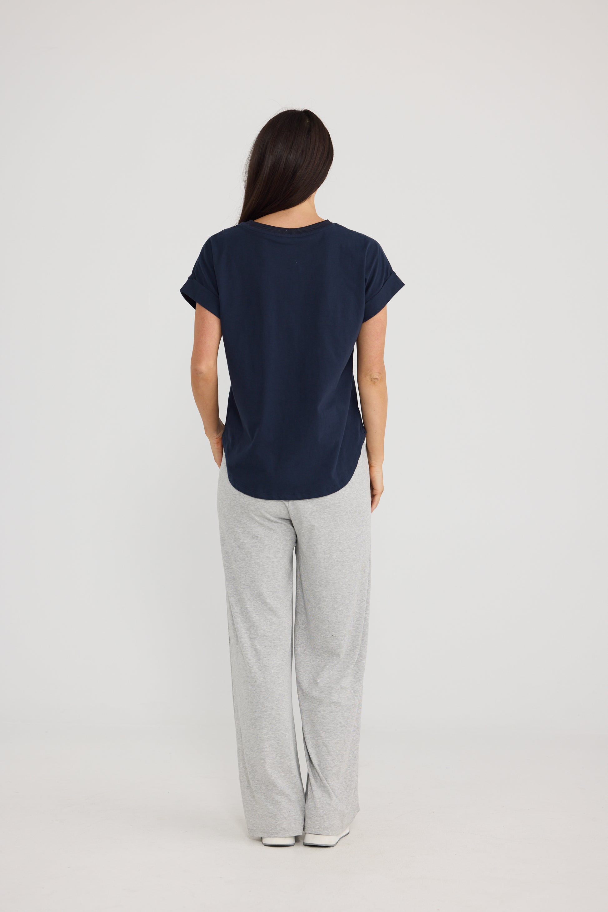 A woman viewed from the back wearing a Brave & True navy blue short-sleeve top and light gray wide-leg pants against a plain light gray studio background.