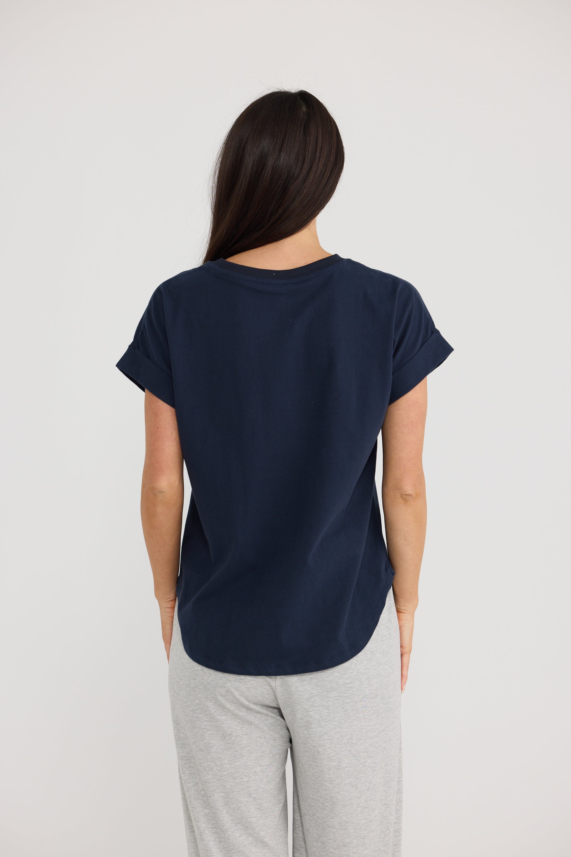 A woman viewed from behind wearing a Brave & True navy blue short-sleeve t-shirt and light gray sweatpants against a plain light gray background.