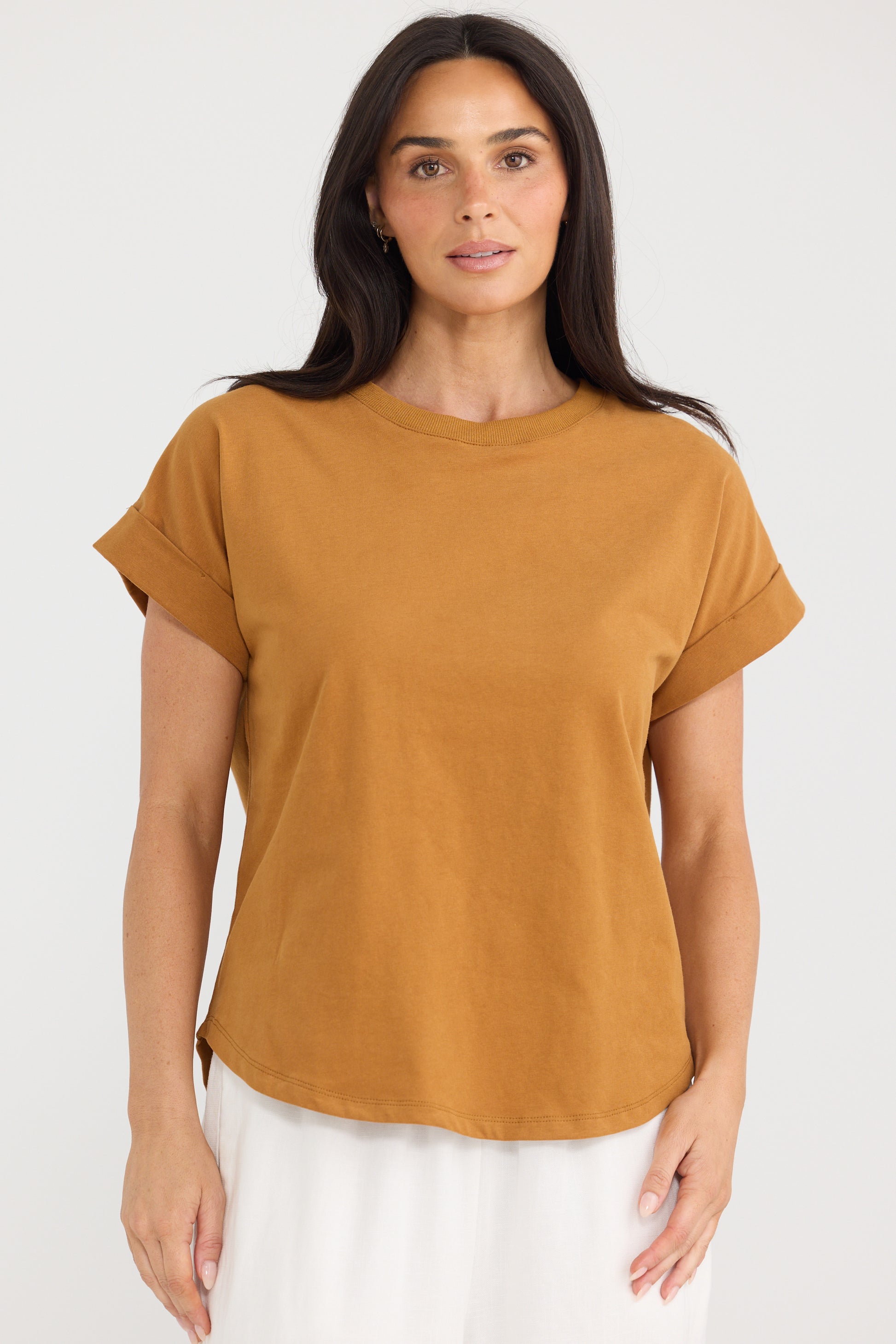 A woman modeling a Brave & True short-sleeve crewneck t-shirt in a warm mustard yellow color, paired with white pants, against a plain light gray background.