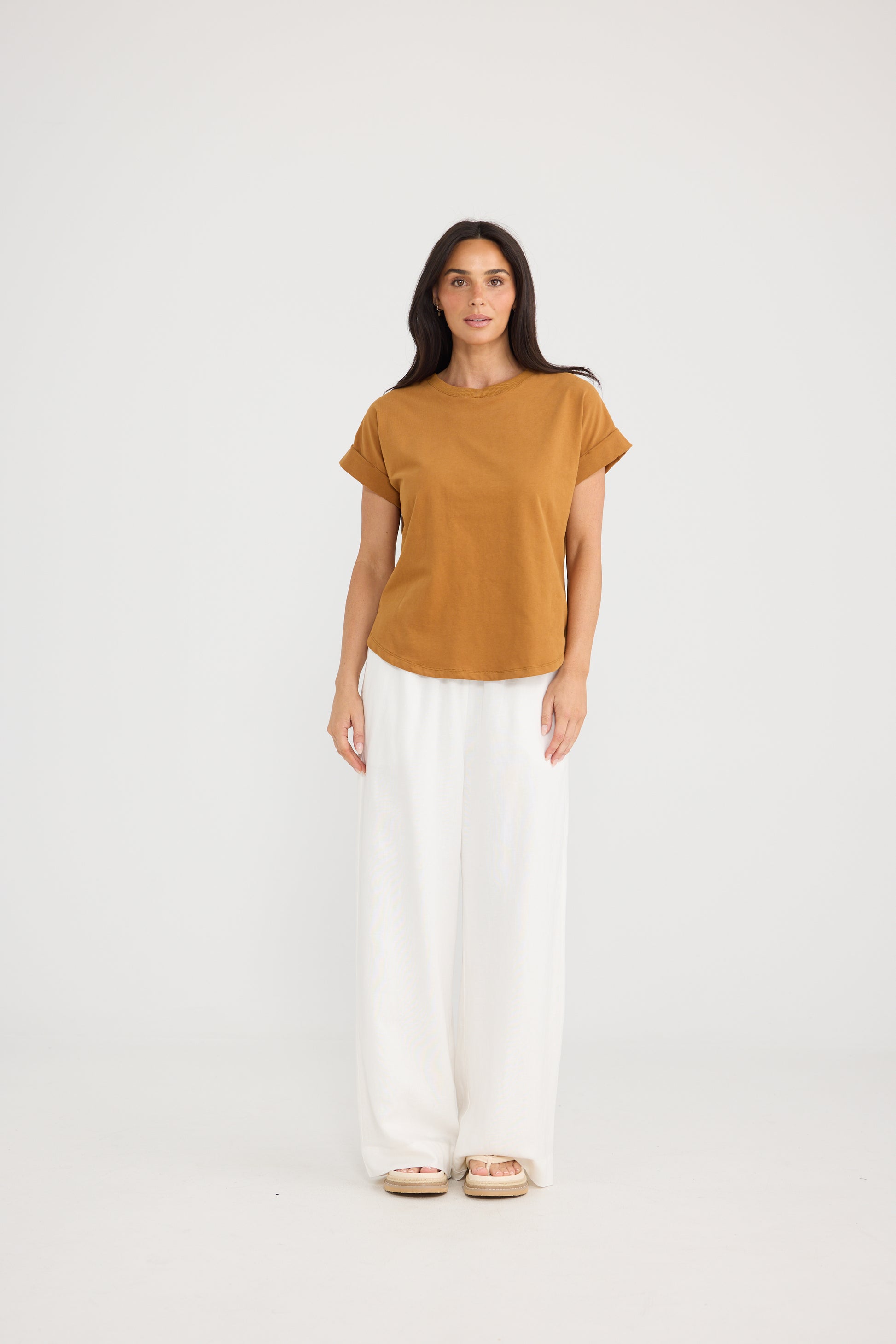 A woman modeling a Brave & True short-sleeve mustard-brown top and white wide-leg pants against a plain light gray studio background.