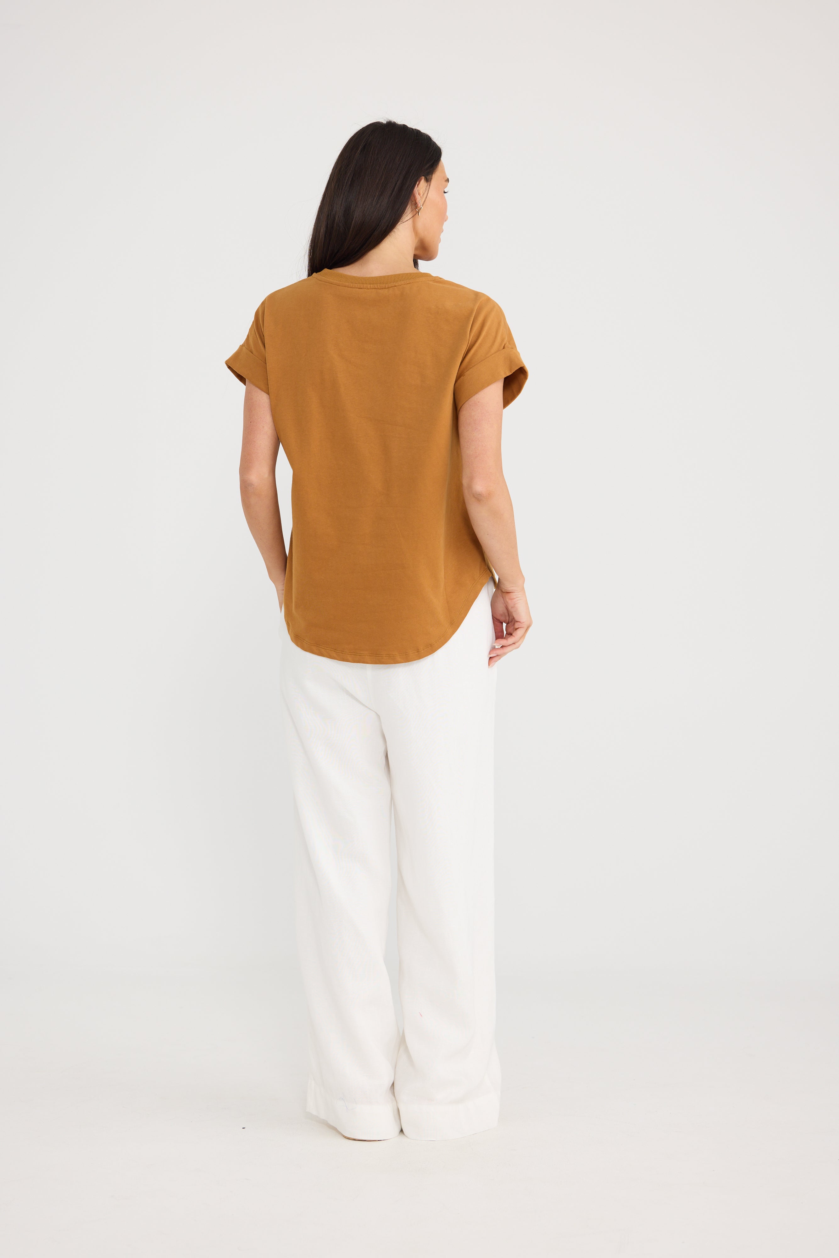 Image of A woman is shown from the back wearing a Brave & True short-sleeve, crew-neck top in a warm camel brown color, paired with white wide-leg pants, against a plain white studio background.