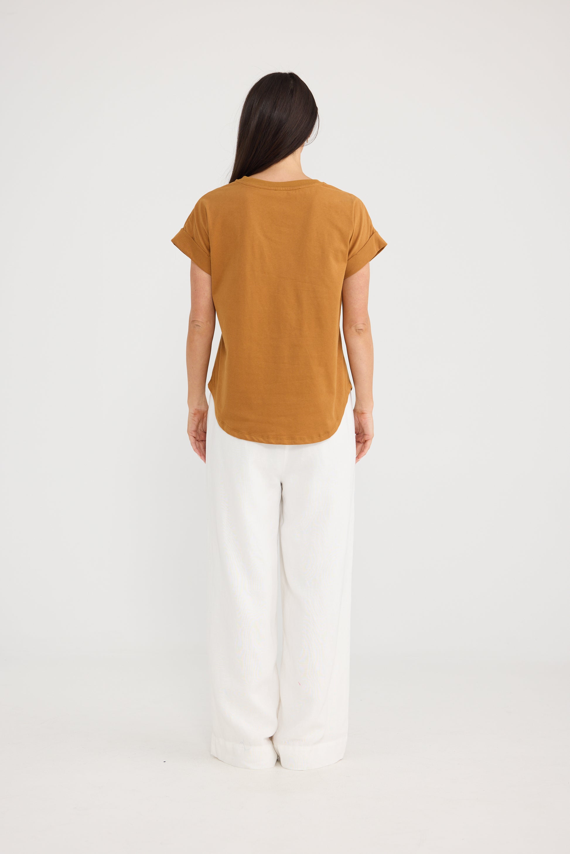 A woman viewed from the back wearing a Brave & True short-sleeve mustard yellow t-shirt and white wide-leg pants, standing against a plain white studio background.