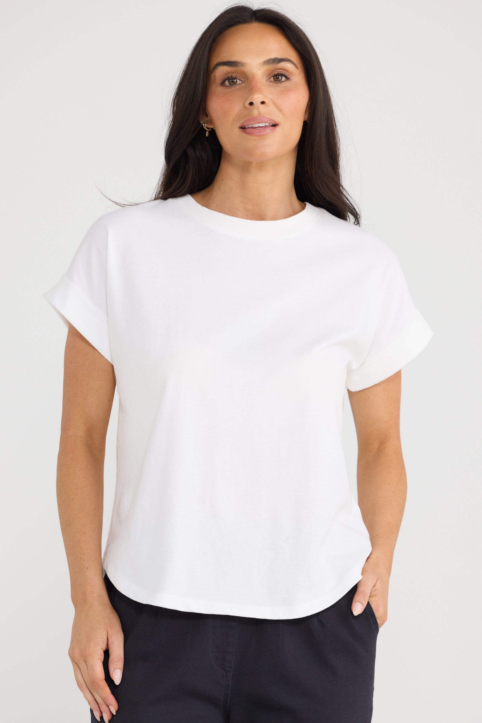 A woman wearing a Brave and True white short-sleeve crewneck top and dark navy pants, standing against a light gray studio background.