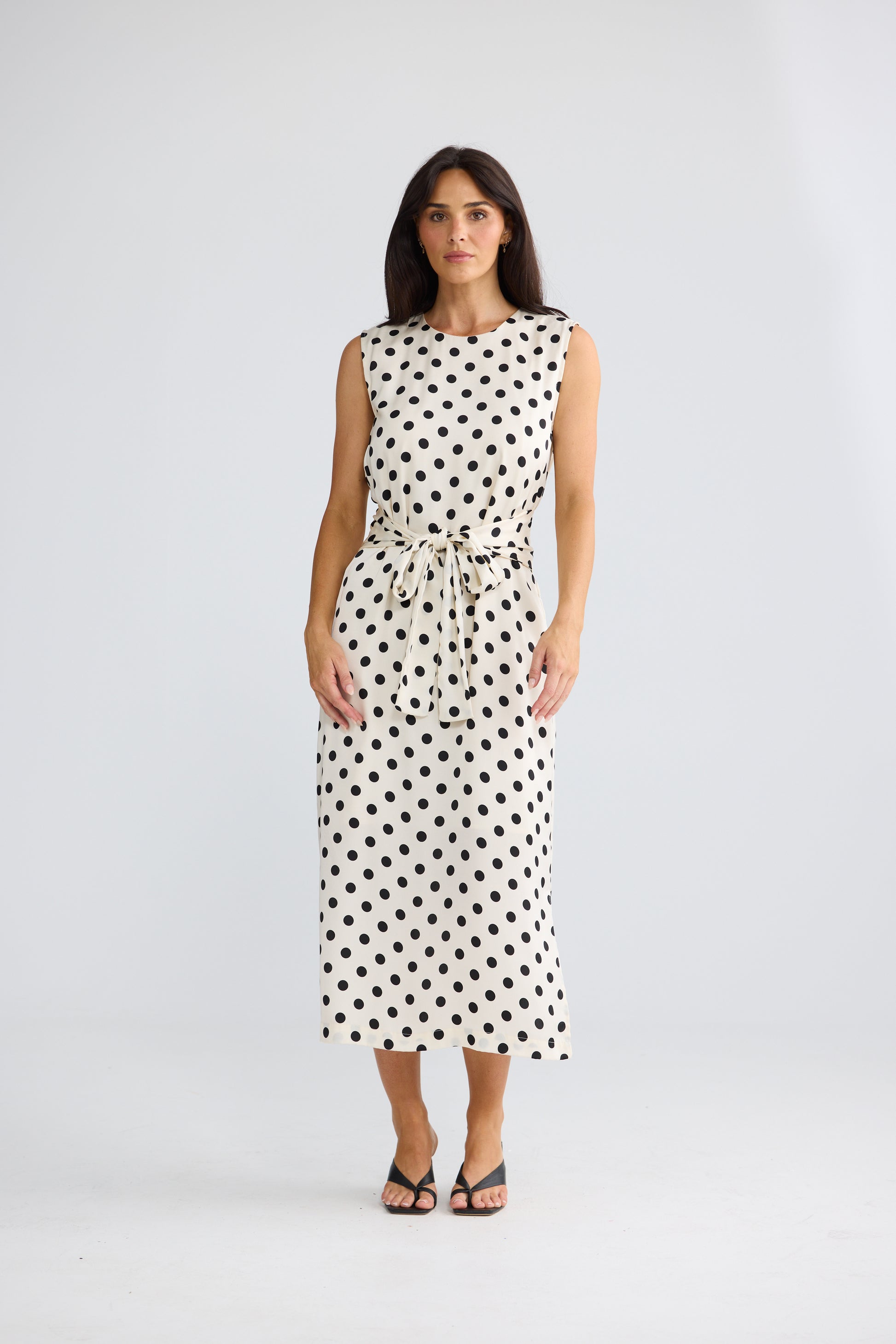 A woman modeling a Brave & True sleeveless midi dress with a black polka dot pattern on a white background, featuring a tie-waist belt and black strappy sandals, set against a plain light gray studio backdrop.