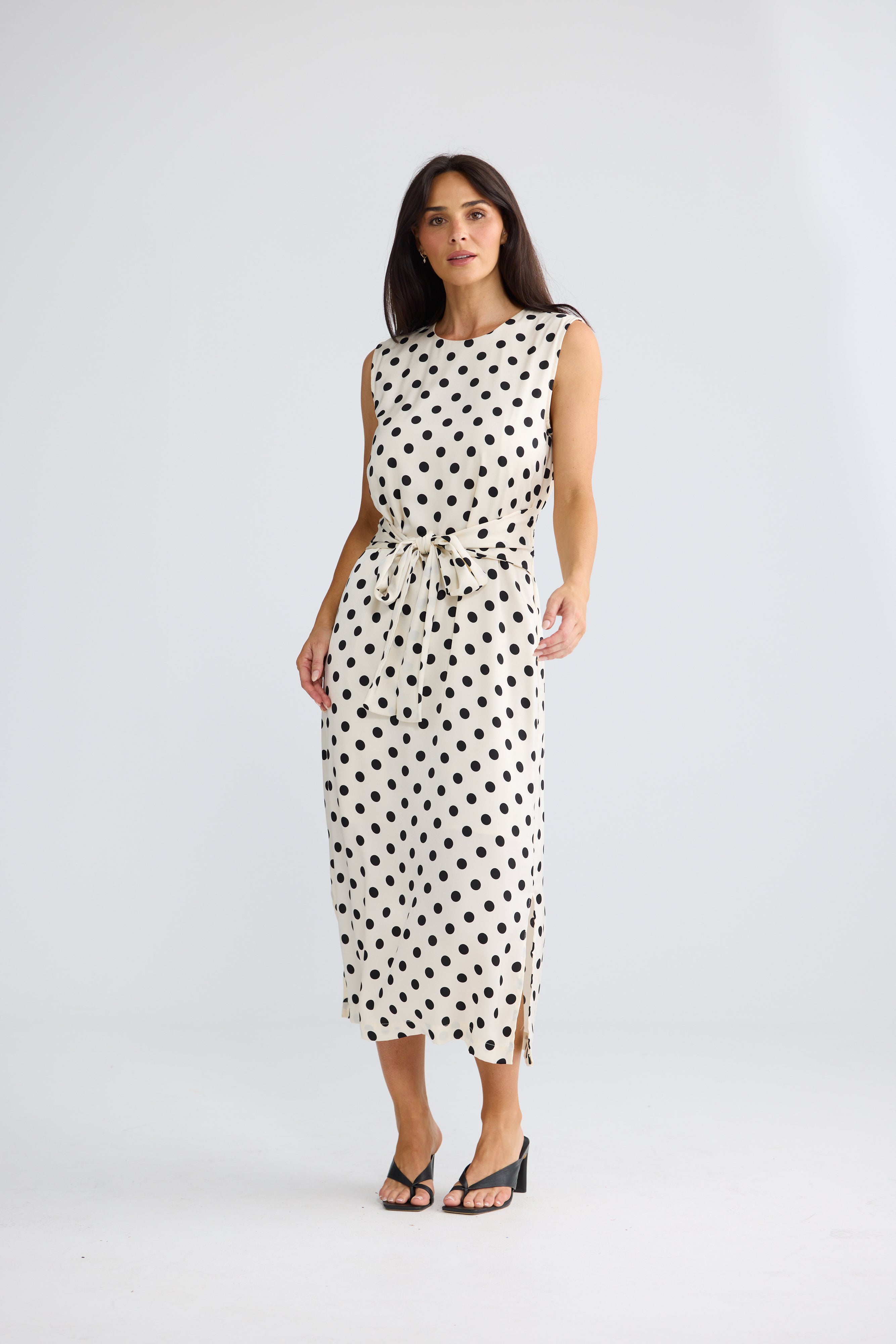 Image of A woman modeling a Brave & True sleeveless midi dress with a black polka dot pattern on a white background, featuring a tie waist and side slits, paired with black sandals against a light gray studio backdrop.