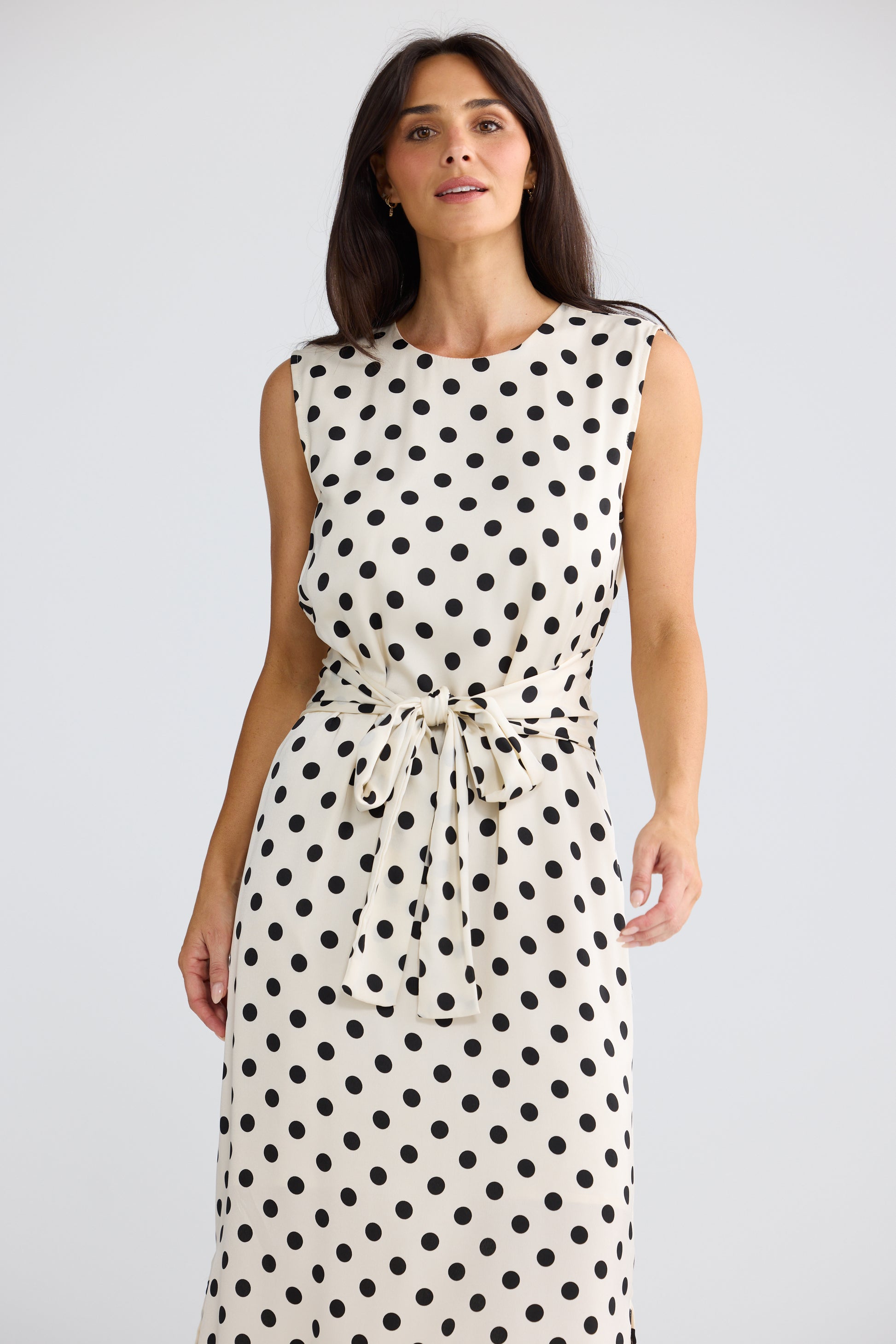 A woman modeling a Brave & True sleeveless midi dress with a black polka dot pattern on a cream background, featuring a tie-waist belt, set against a plain light gray studio background.