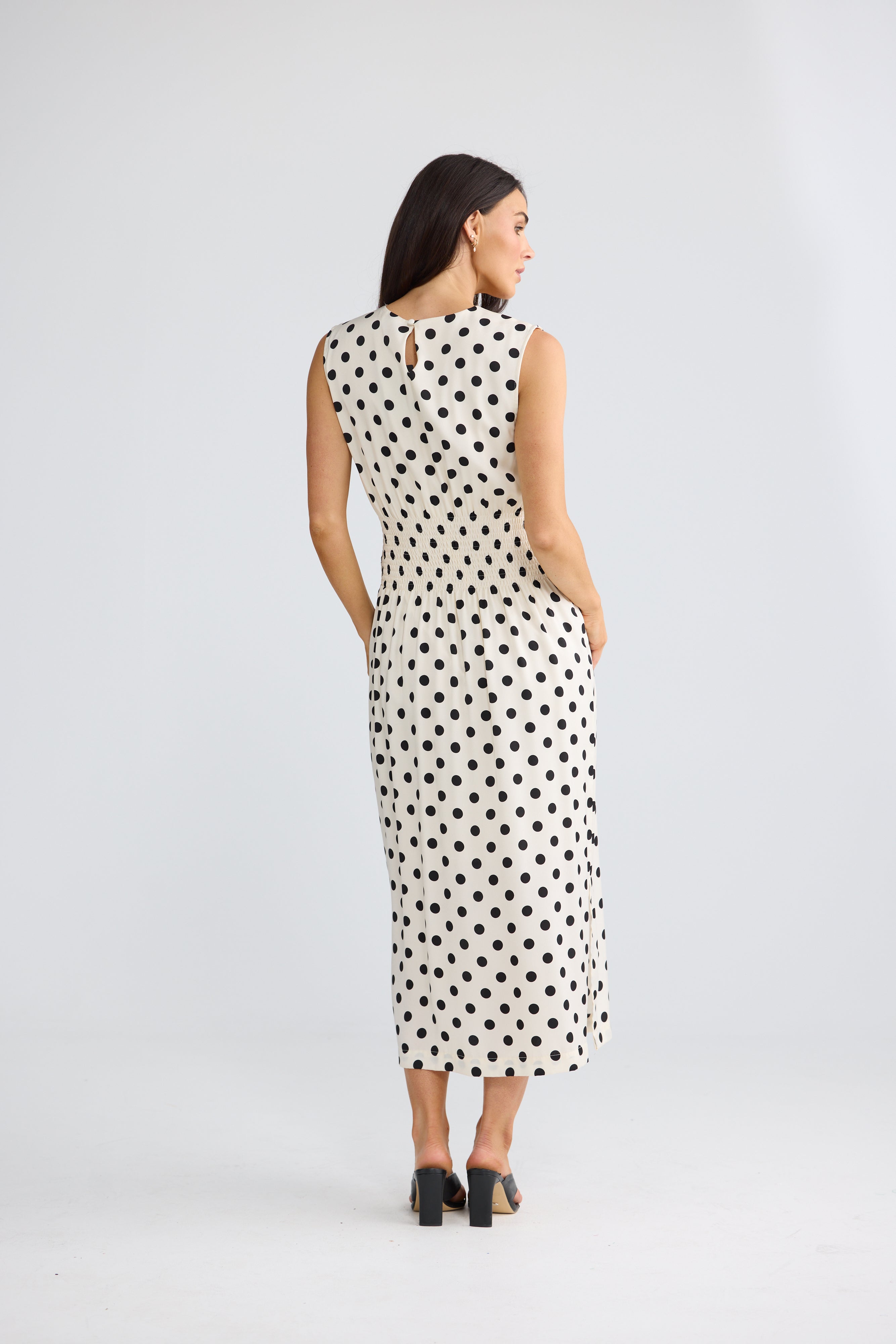 Image of A woman viewed from the back wearing a Brave & True sleeveless midi dress with a black polka dot pattern on a white background, paired with black high heels, against a light gray studio backdrop.