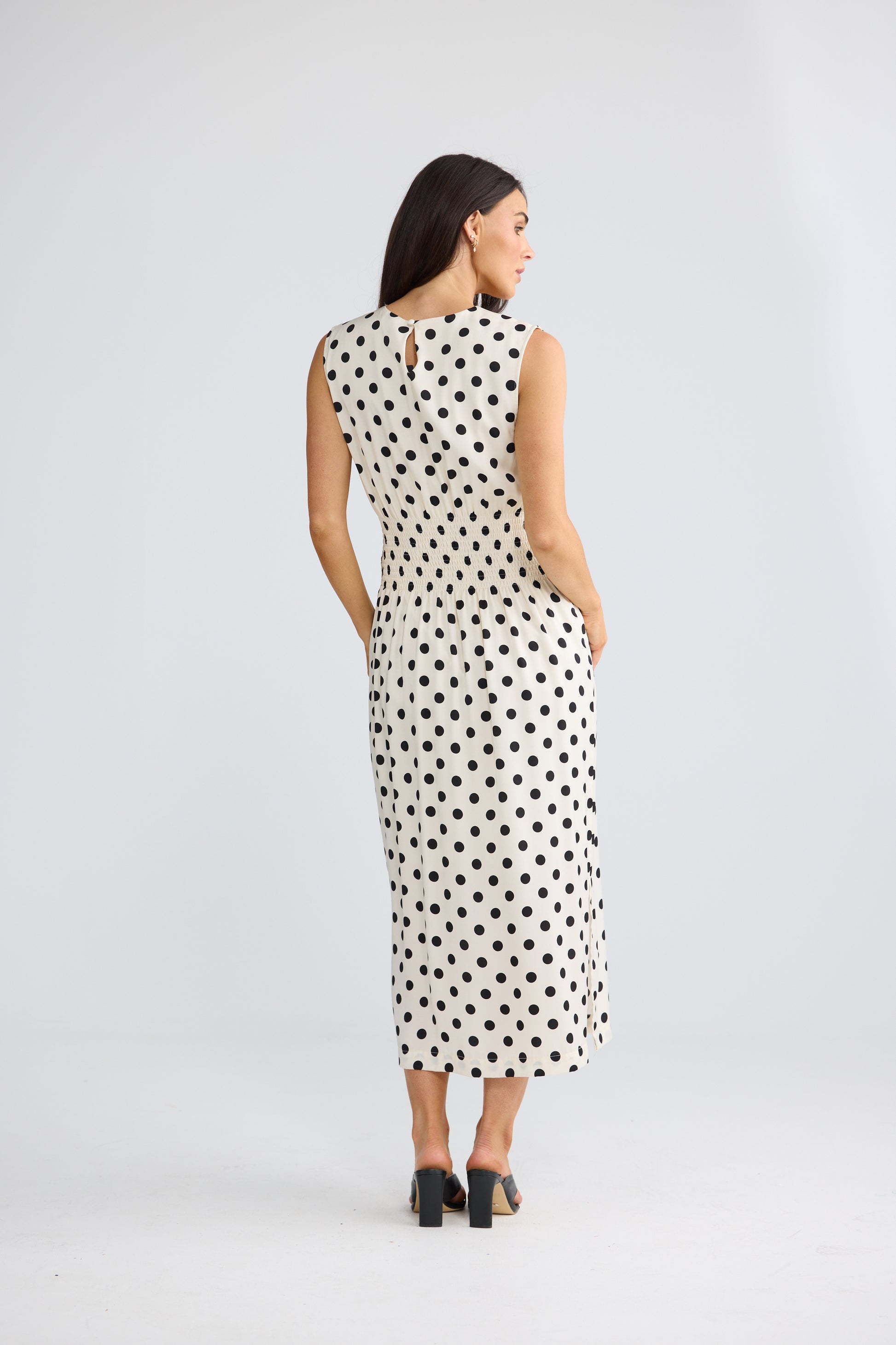A woman viewed from the back wearing a Brave & True sleeveless midi dress with a black polka dot pattern on a white background, paired with black high heels, against a light gray studio backdrop.