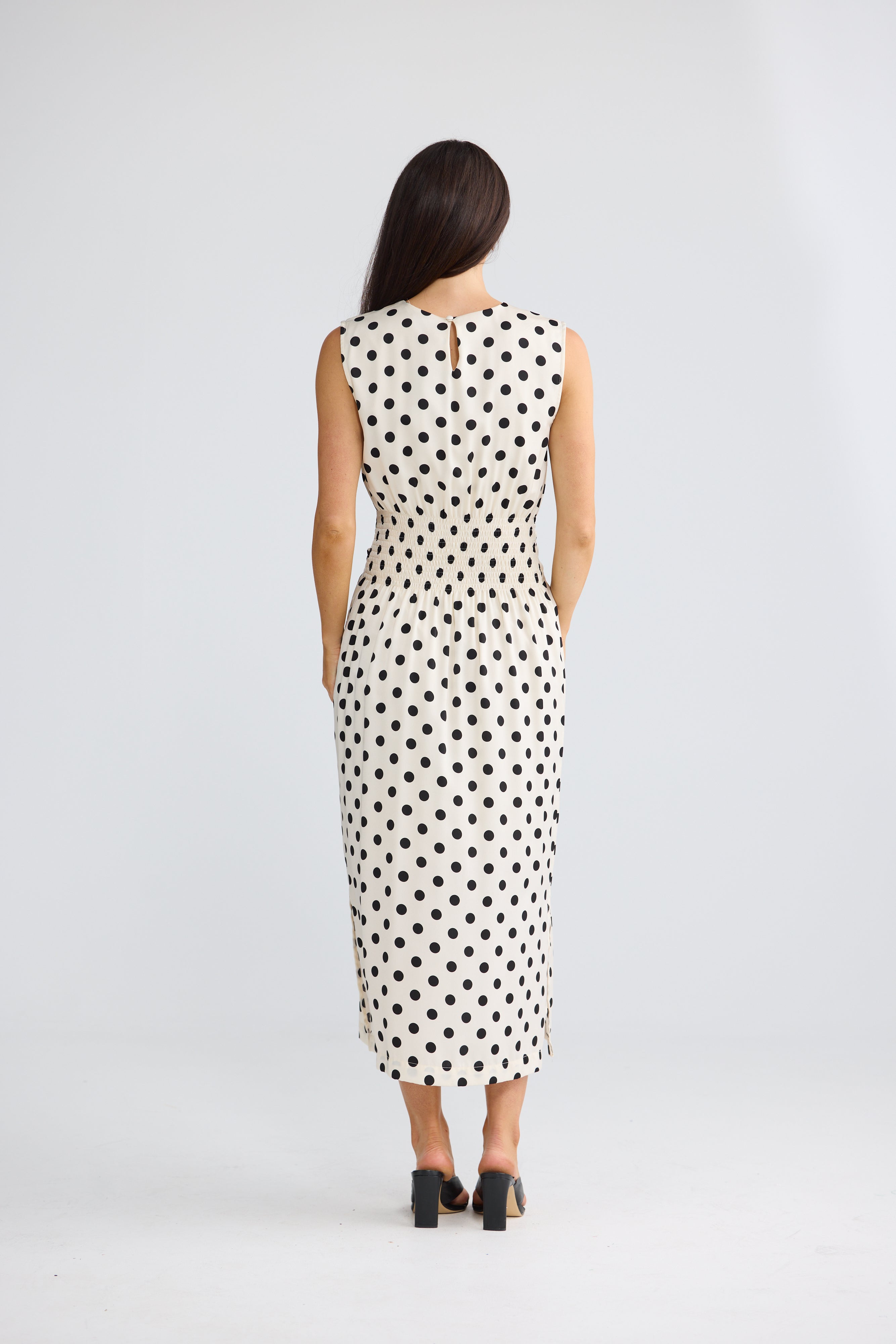 Image of A woman viewed from behind wearing a Brave & True sleeveless midi dress with a black polka dot pattern on a white background, paired with black high heels, against a light gray studio backdrop.