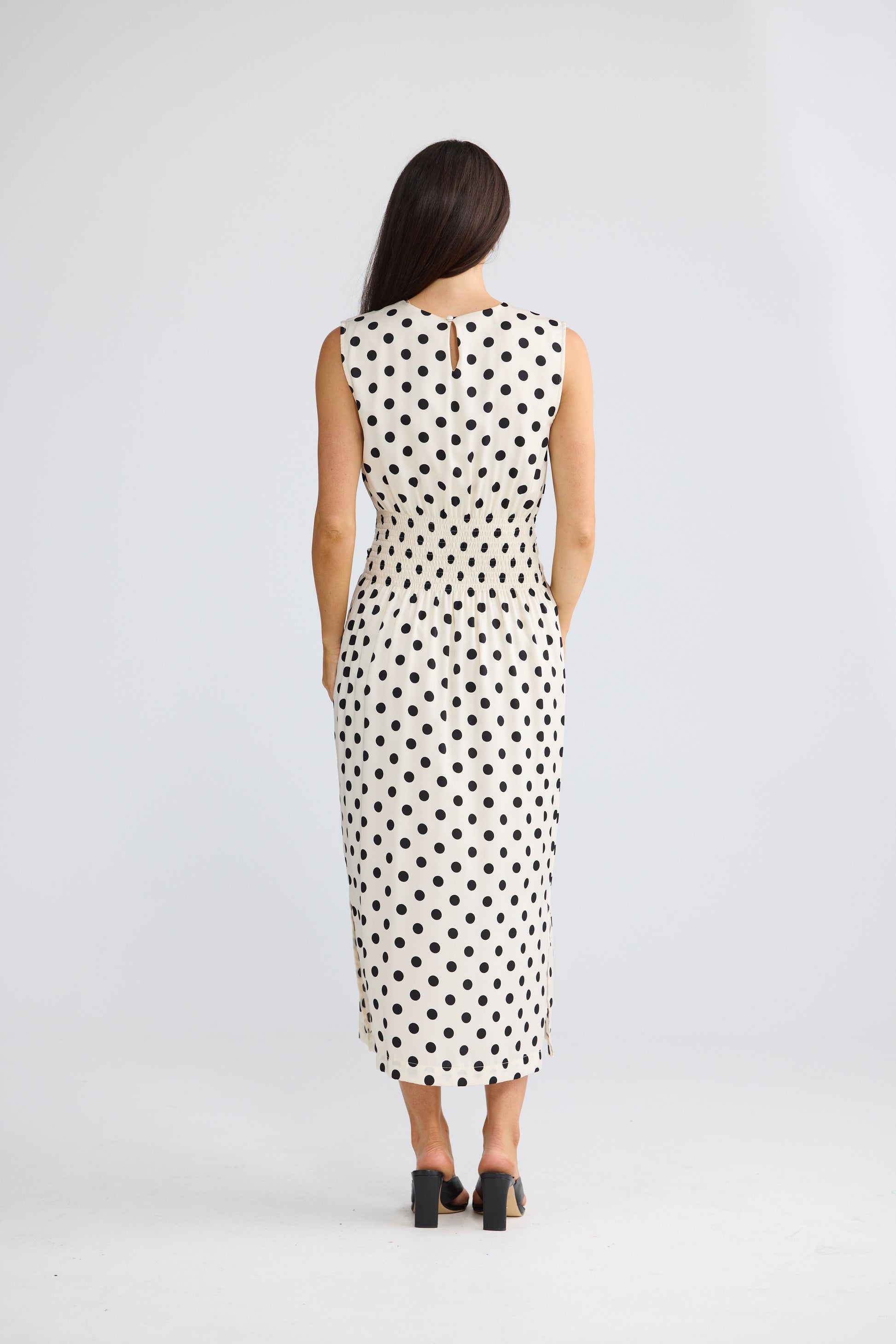 A woman viewed from behind wearing a Brave & True sleeveless midi dress with a black polka dot pattern on a white background, paired with black high heels, against a light gray studio backdrop.