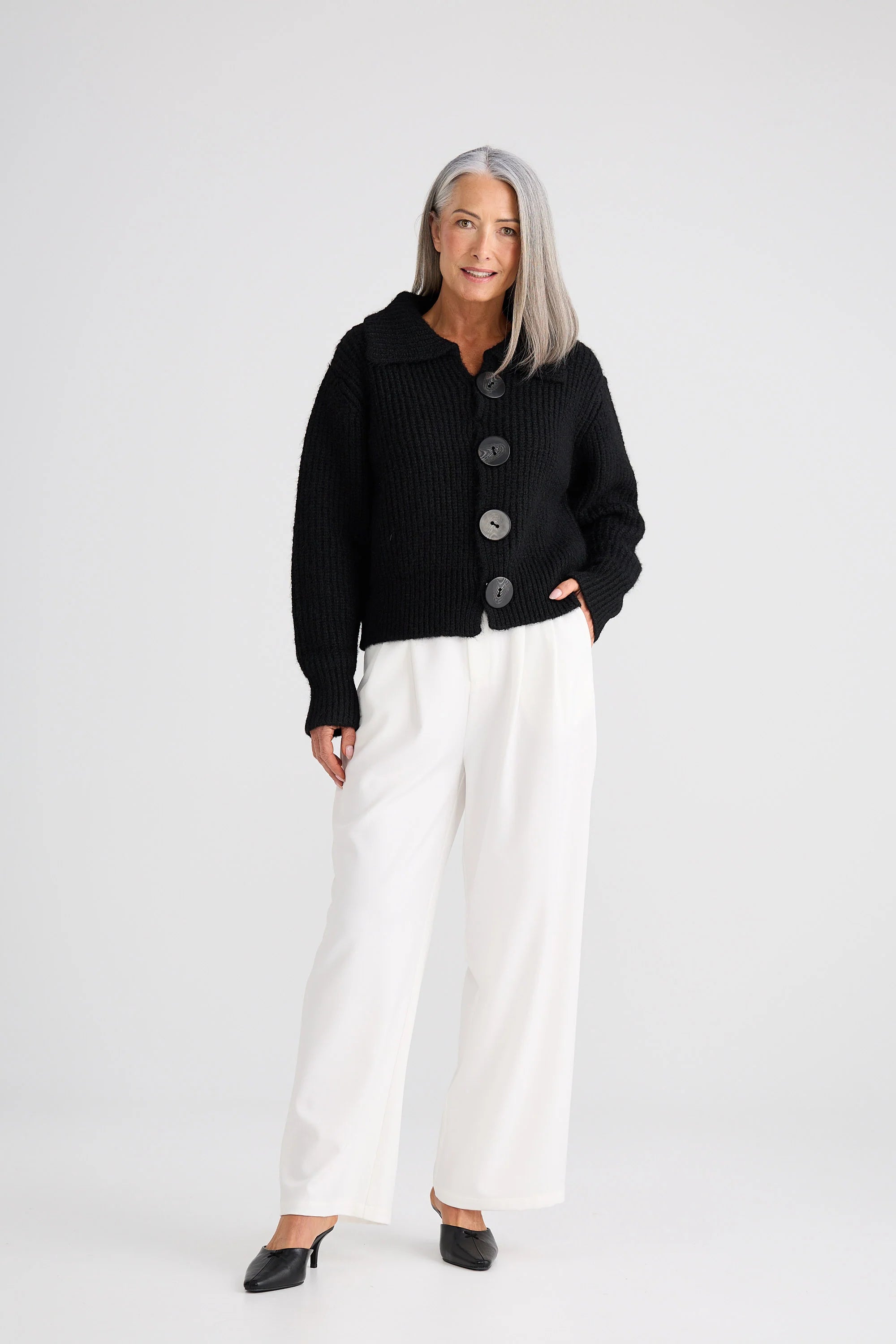 Image of Woman wearing a Brave and True black cardigan with buttons and white pants on a white background