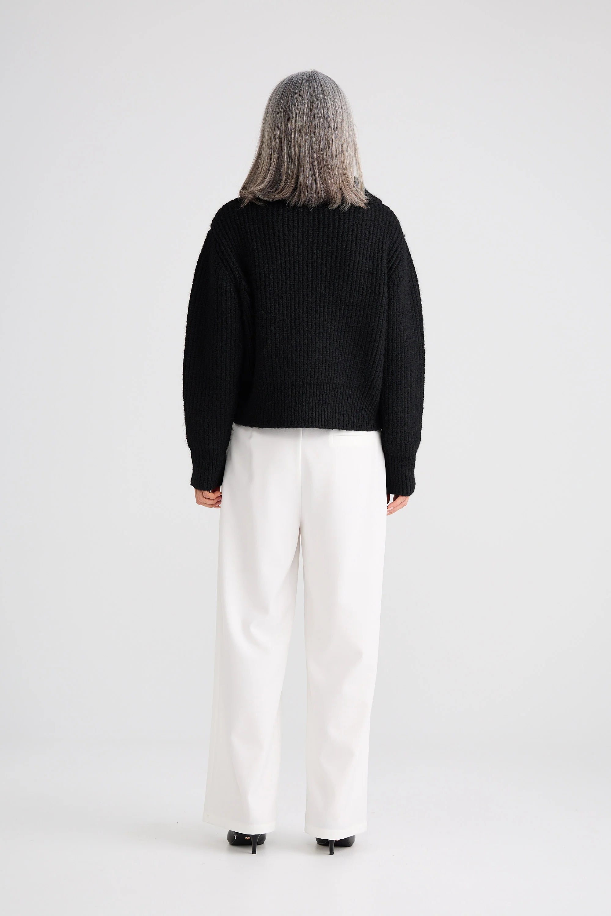 Image of Woman wearing a Brave and True black sweater and white pants on a plain background
