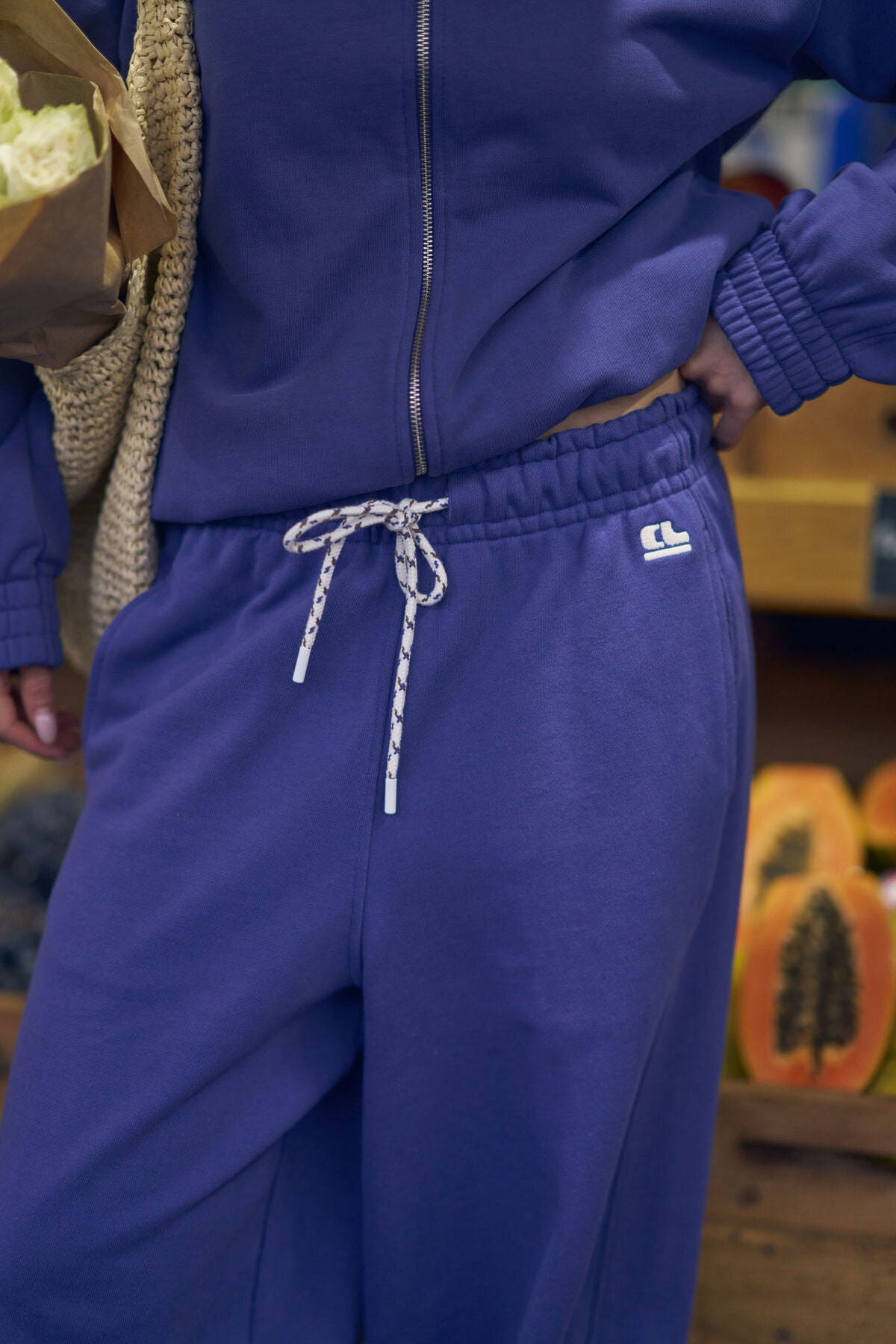 Person wearing a blue tracksuit with a brand logo, holding a woven bag.