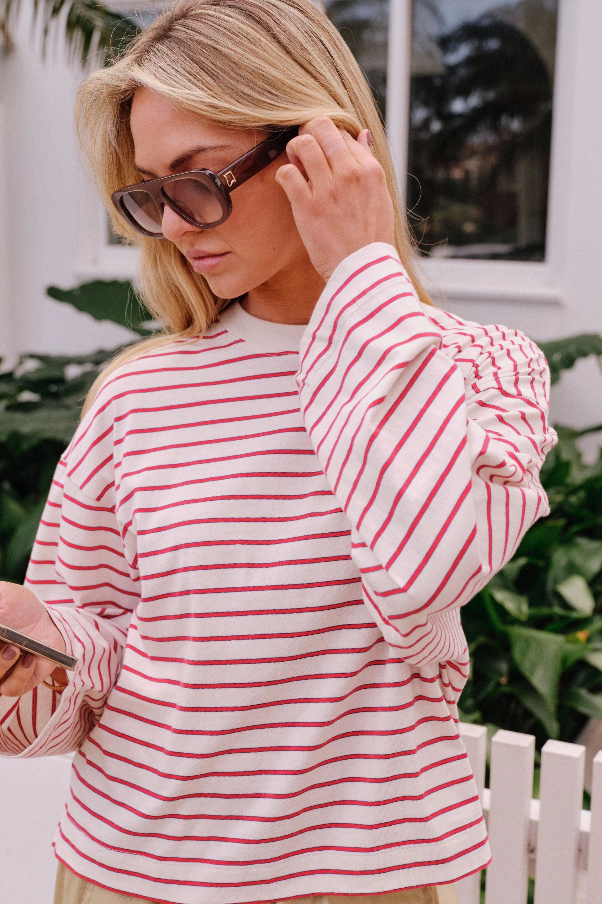 Image of A woman wearing a Ceres Life pink and white striped long-sleeve crop top and beige wide-leg pants, leaning against a white picket fence outdoors.
