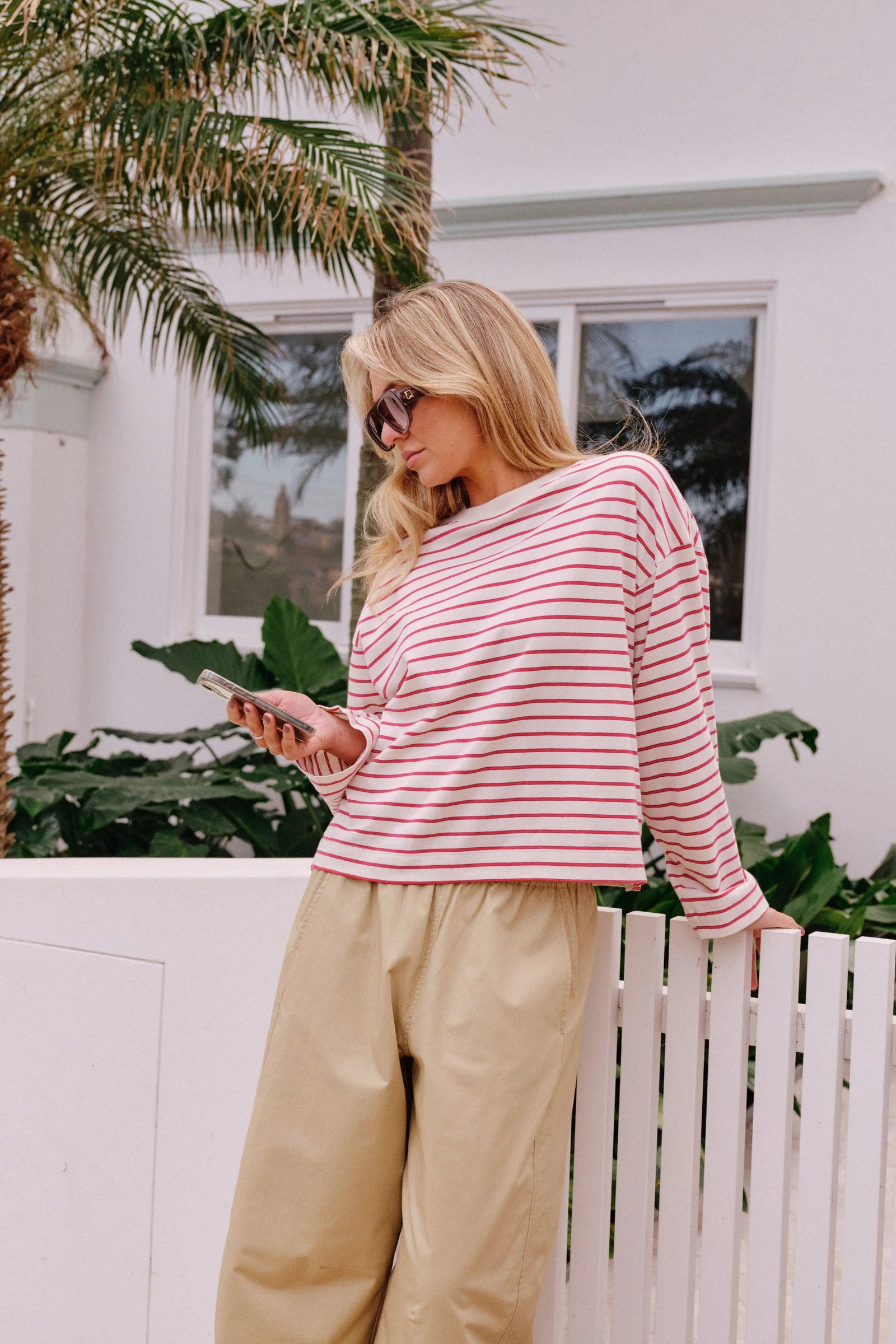 Image of A woman wearing a Ceres Life pink and white striped long-sleeve crop top and beige wide-leg pants, leaning against a white picket fence outdoors.