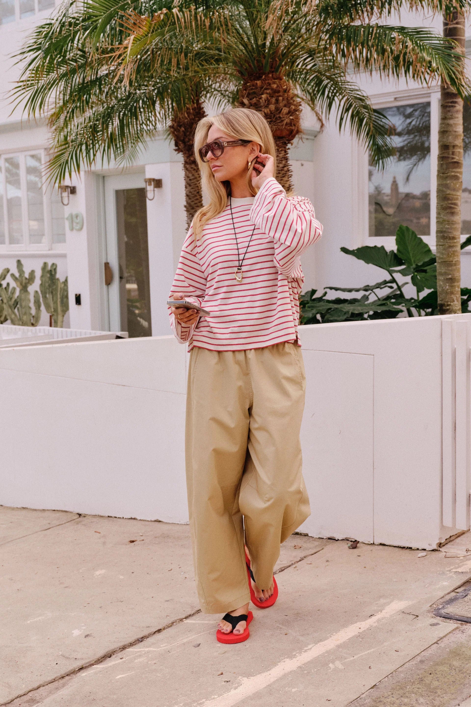 Image of A woman wearing a Ceres Life pink and white striped long-sleeve crop top and beige wide-leg pants, leaning against a white picket fence outdoors.