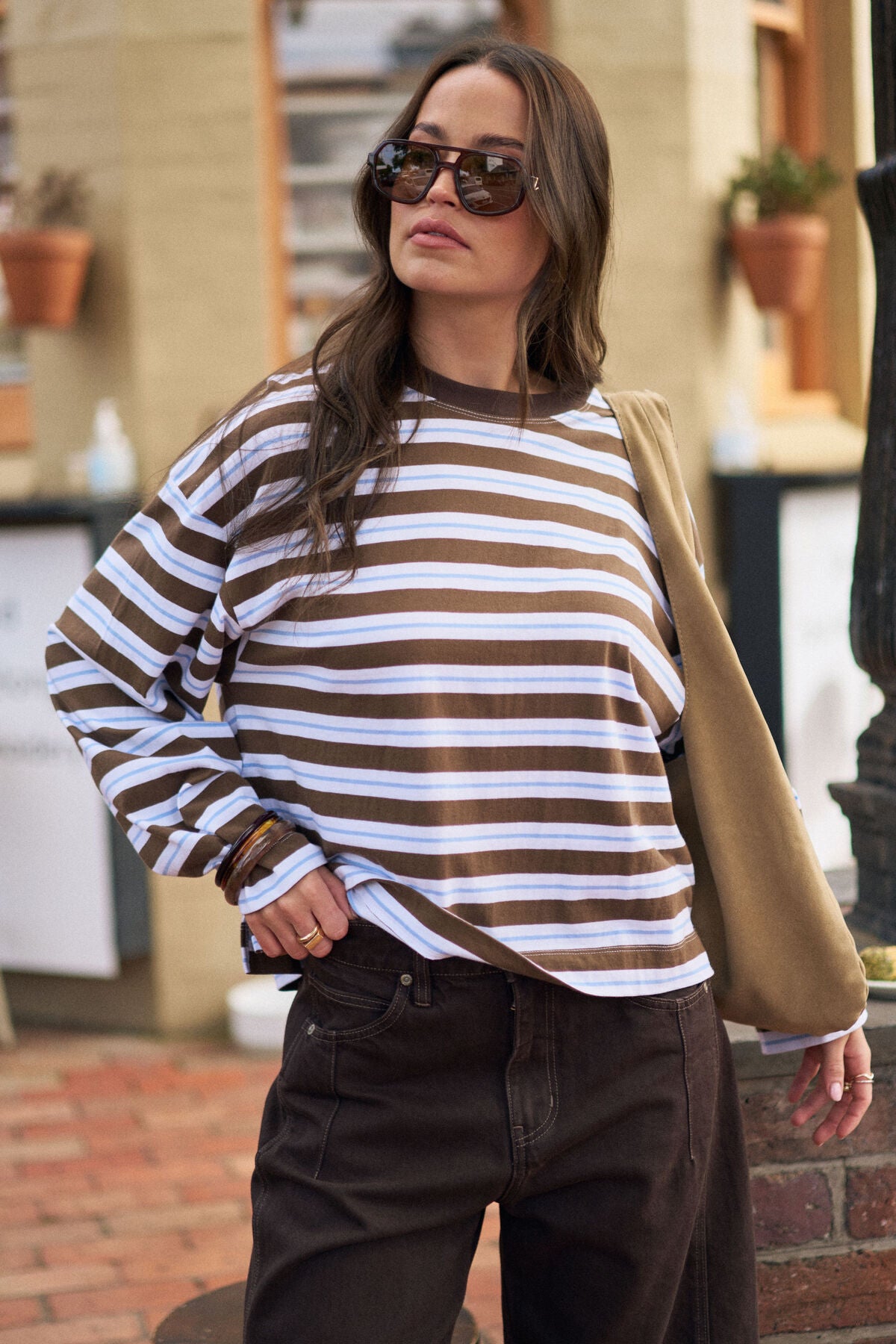 Woman wearing a Ceres Life brown and white striped sweater with sunglasses and a beige bag outdoors.